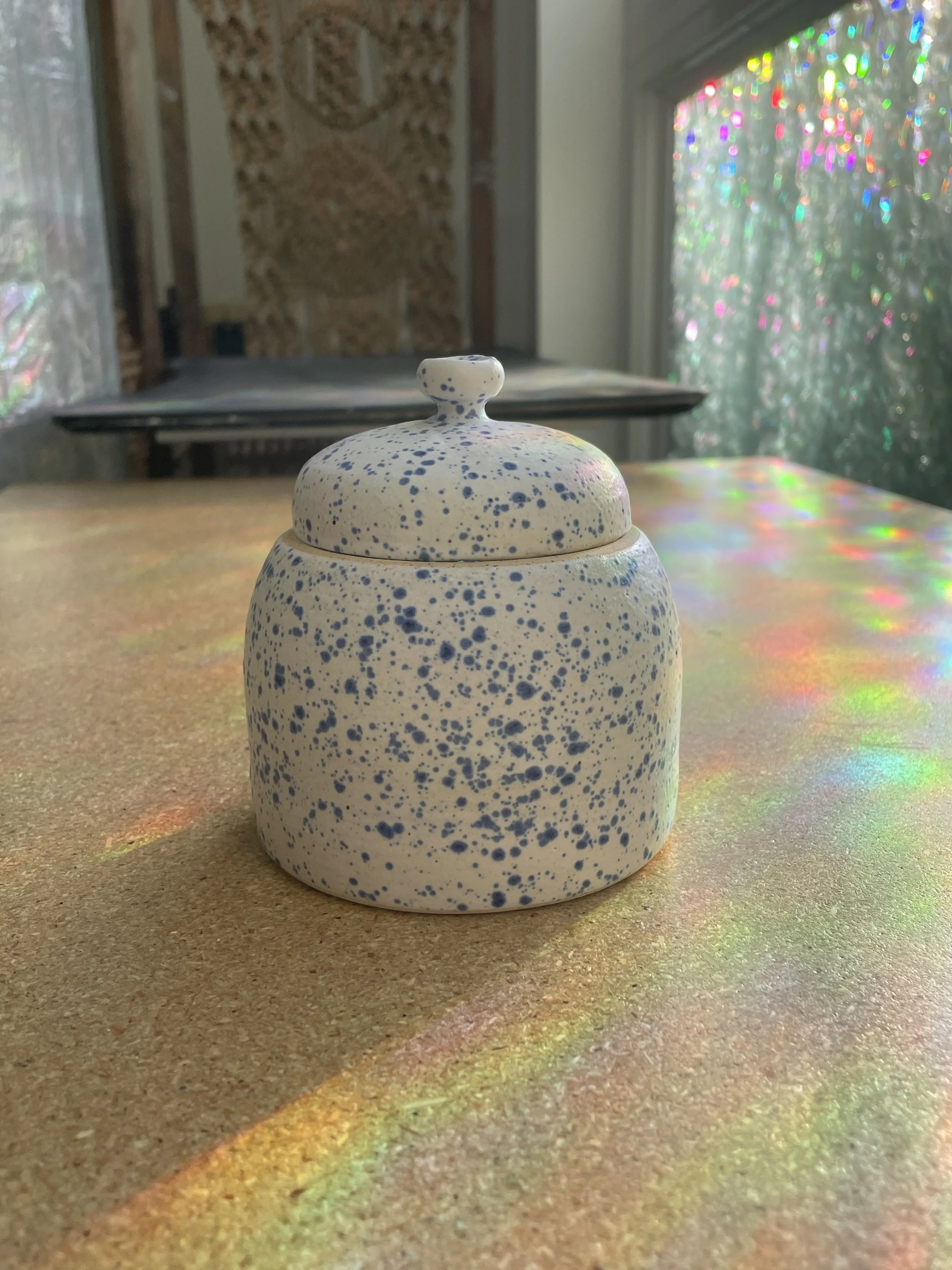 A ceramic jar with a matching lid, speckled blue and white design, near a window with colorful light reflections.
