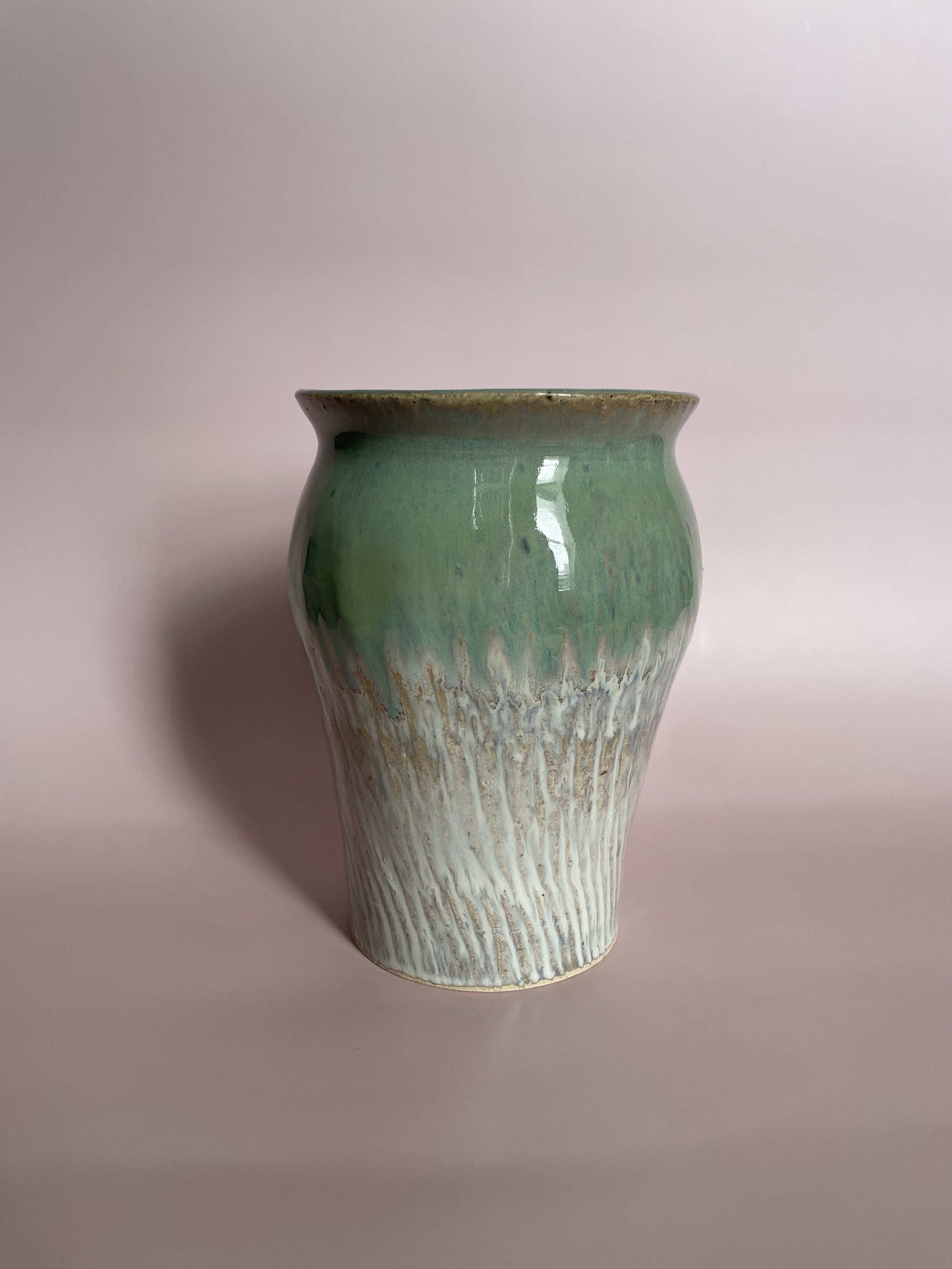 A ceramic vase with a green upper half and a textured white lower half, placed against a light pink background.