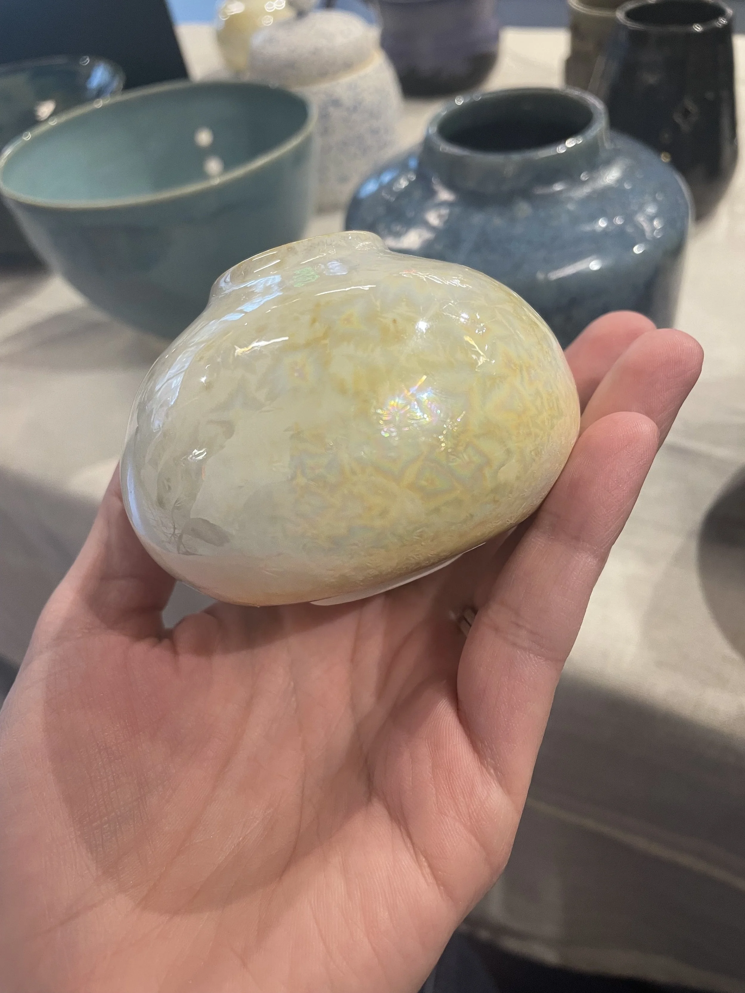 A hand holding a glossy, iridescent mini vase with several ceramic vases and bowls in shades of blue, white, and black in the background.