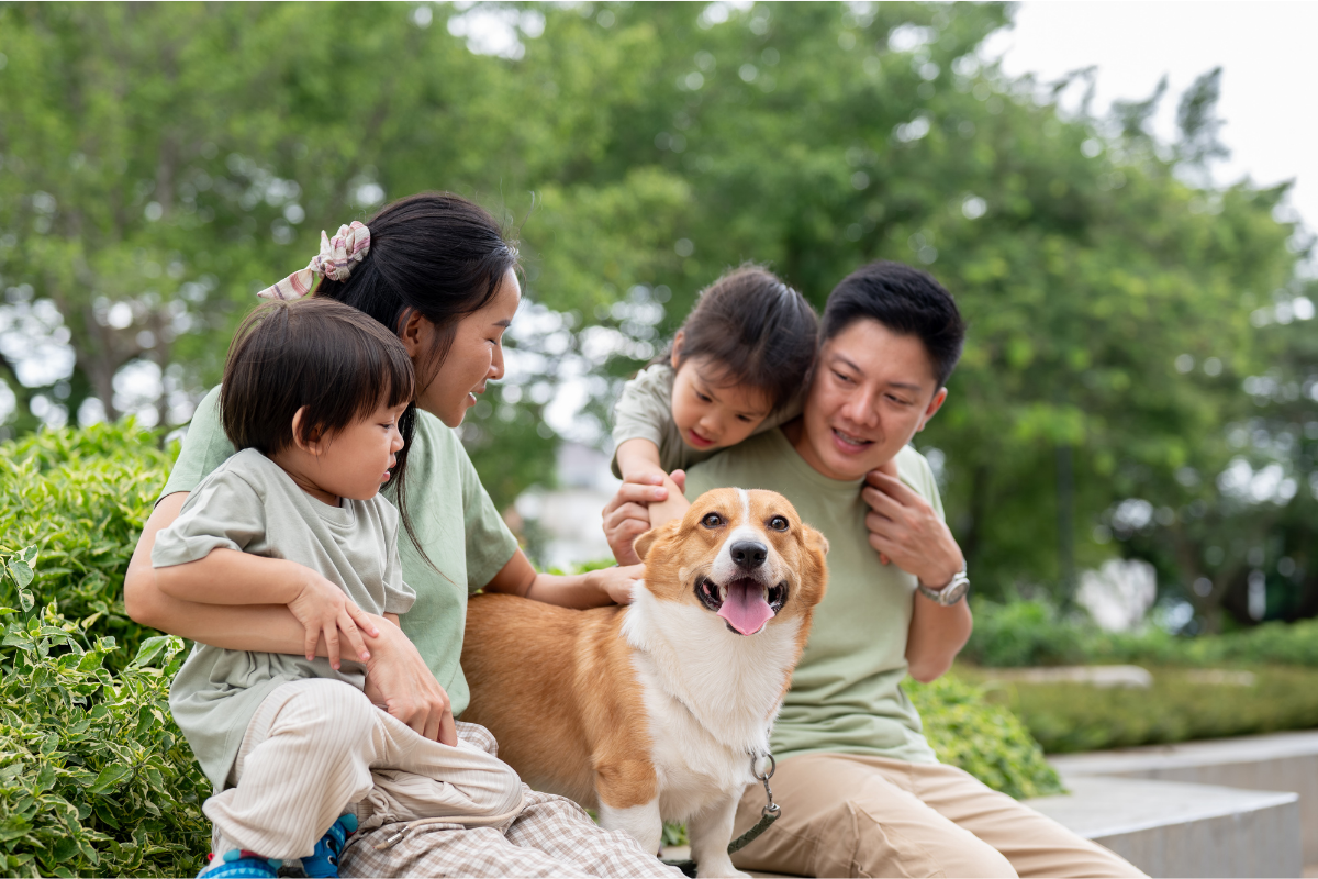 Family with two children and a dog sitting outdoors on a park bench with green trees in the background.