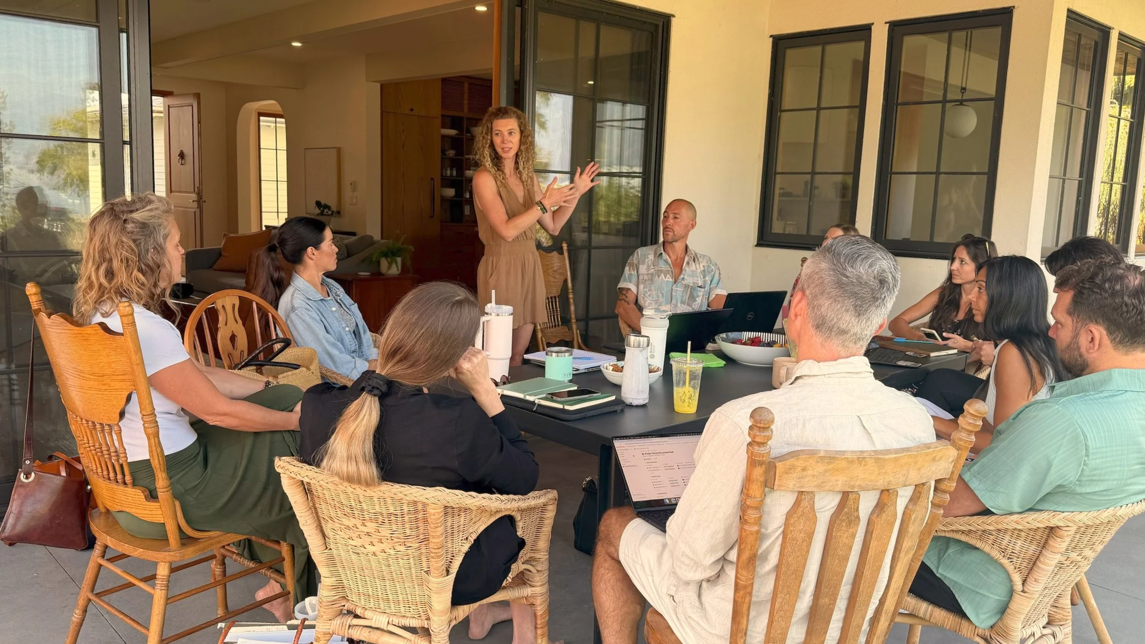 A woman is standing and speaking to a group of people sitting at a large table on an outdoor patio. The group listens attentively, with some taking notes or using laptops.