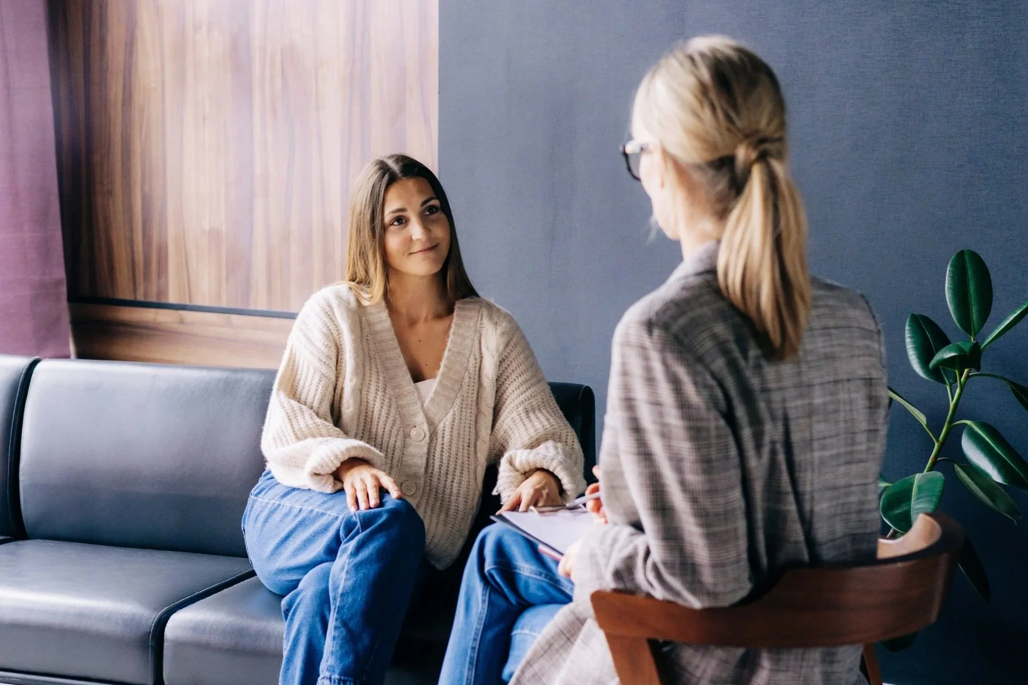 A woman in a beige cardigan speaking with a therapist in a gray plaid jacket during a session in a modern office.