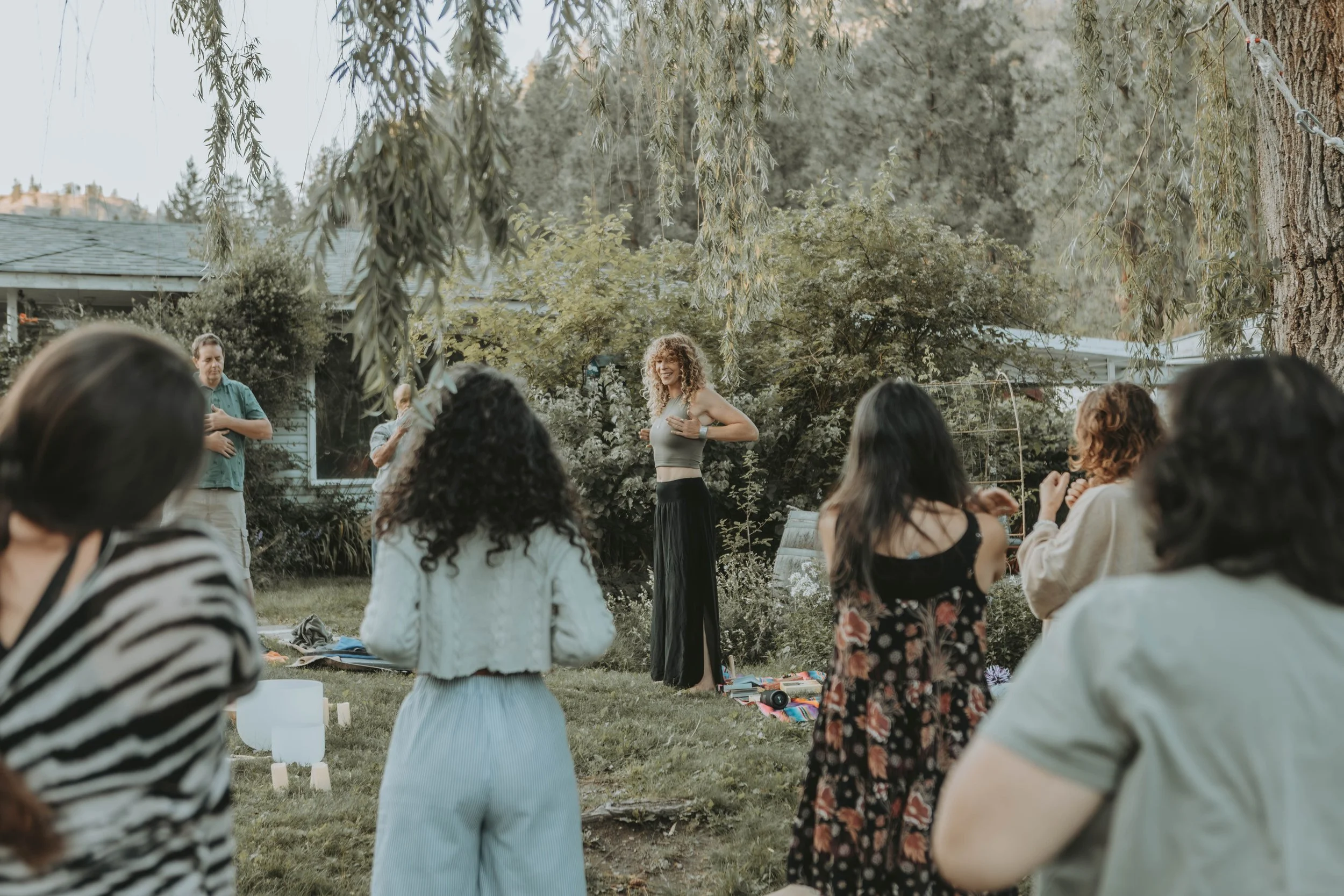 A woman with curly hair stands in the center of a backyard gathering, speaking to a group of women and two men, surrounded by trees and a house, with sunlight filtering through the leaves.