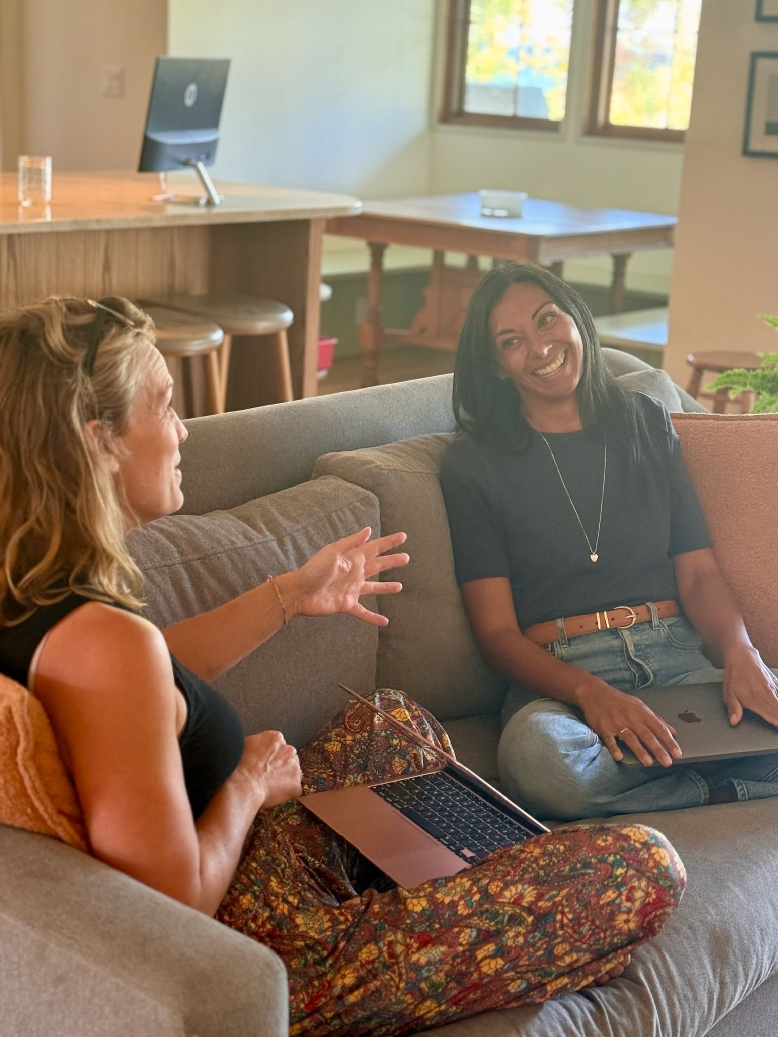 Two women sitting on a sofa and talking in a cozy, well-lit room with windows, a desk, and a computer in the background.