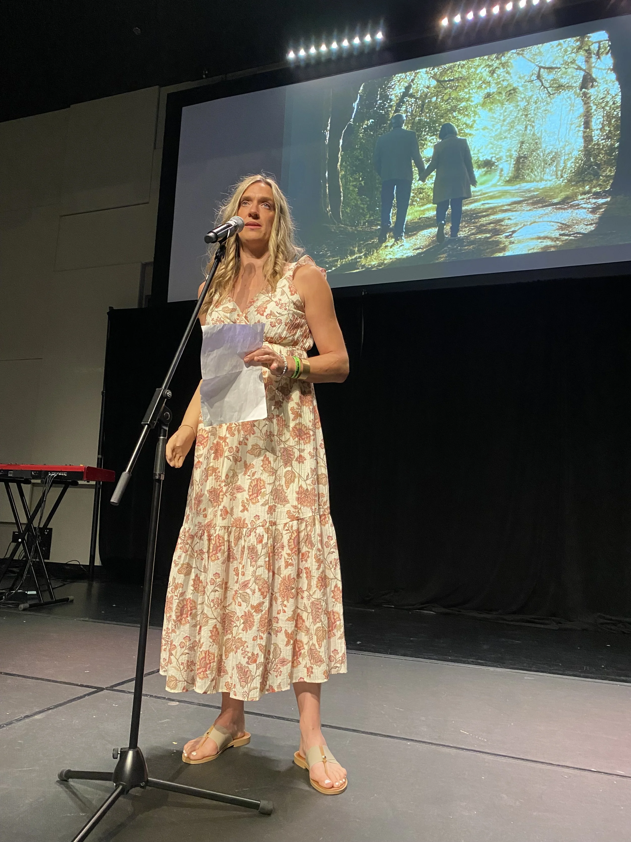 A woman with long blonde hair stands on stage, speaking into a microphone, holding a piece of paper. She wears a long, floral dress and sandals. Behind her, a large screen displays an image of a couple walking hand in hand down a forested path.