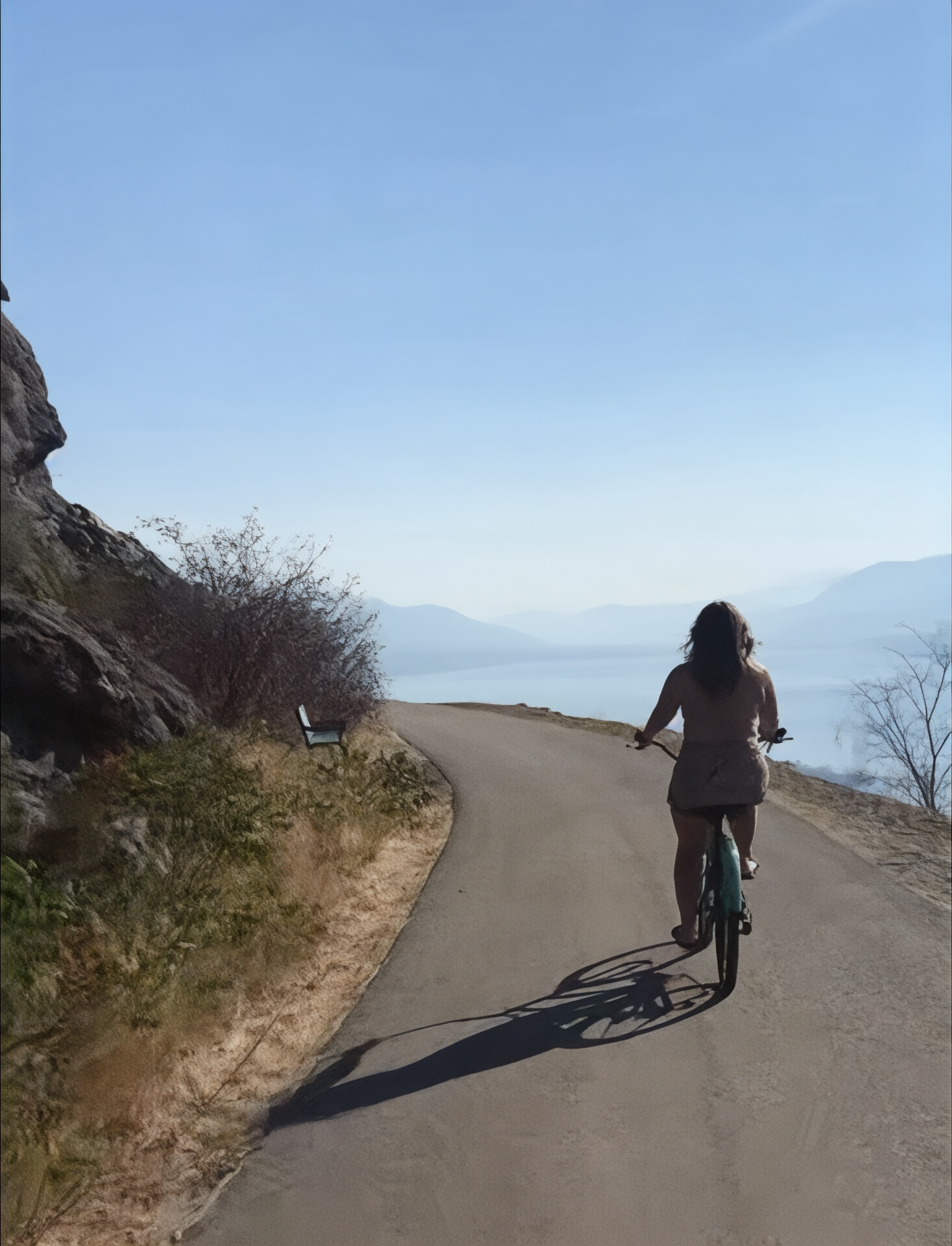 A woman riding a bicycle on a winding road by a lake, with mountains in the background and a clear blue sky.