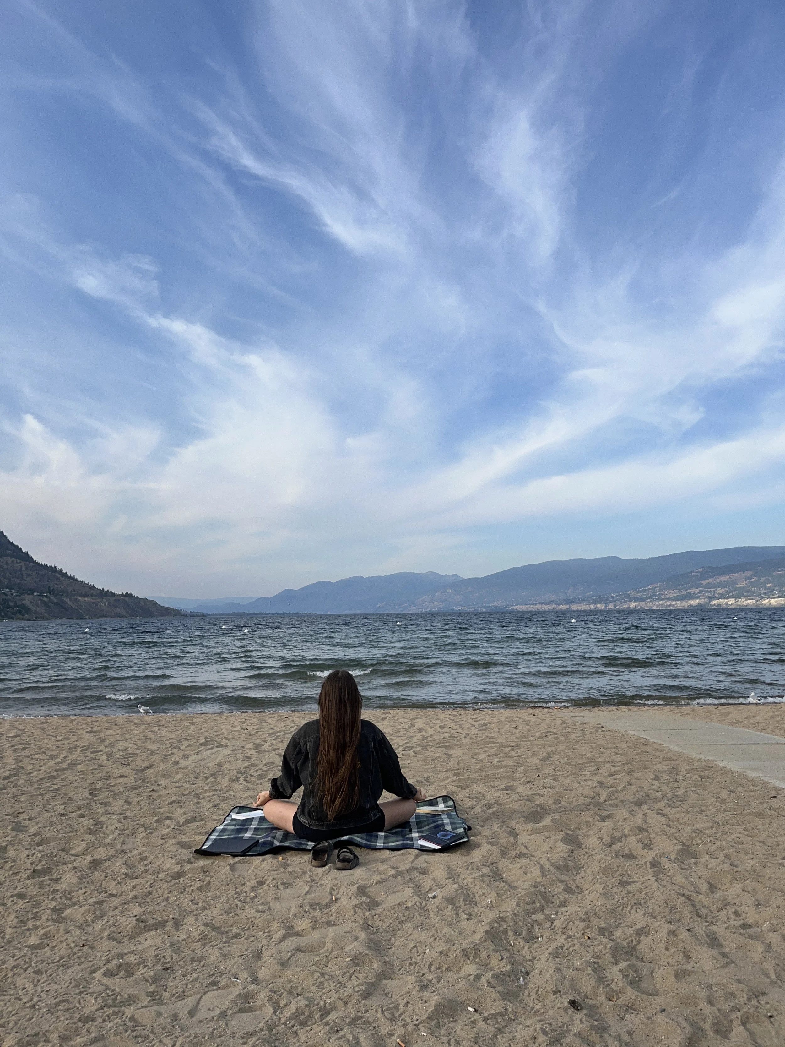 A woman with long brown hair sits cross-legged on a plaid blanket on the sandy beach, facing the water, with mountains in the background and blue sky with wispy clouds overhead.