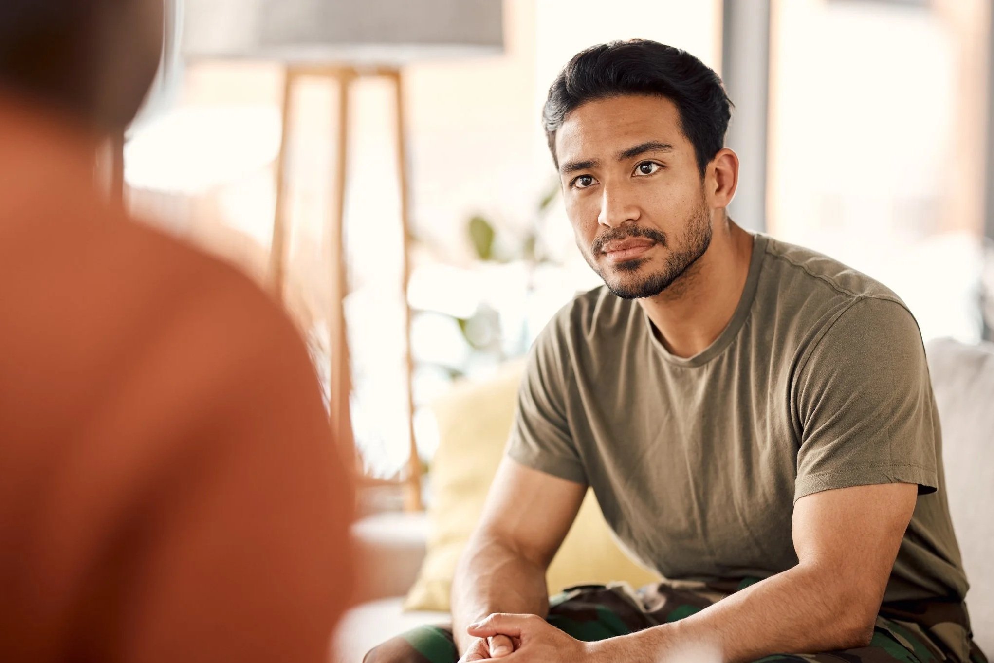 A young man with dark hair and a beard sitting on a beige sofa, listening attentively to someone out of focus in the foreground, with a neutral expression in a well-lit living room.