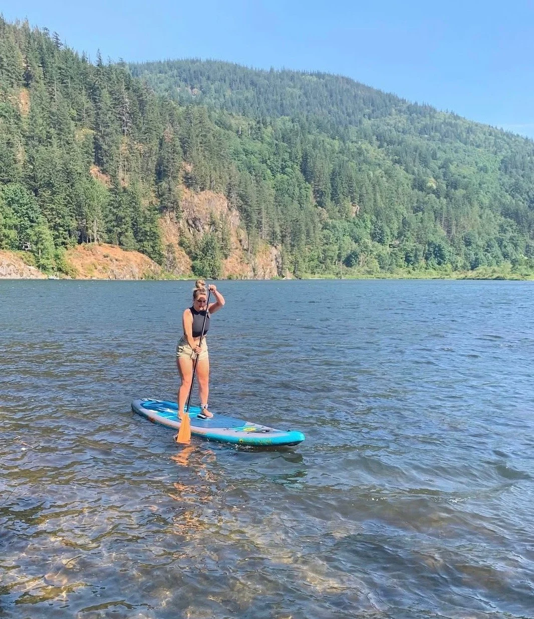 A woman stands on a paddleboard on a lake surrounded by mountains and dense green forest, wearing sunglasses, a black tank top, and shorts, holding a paddle.