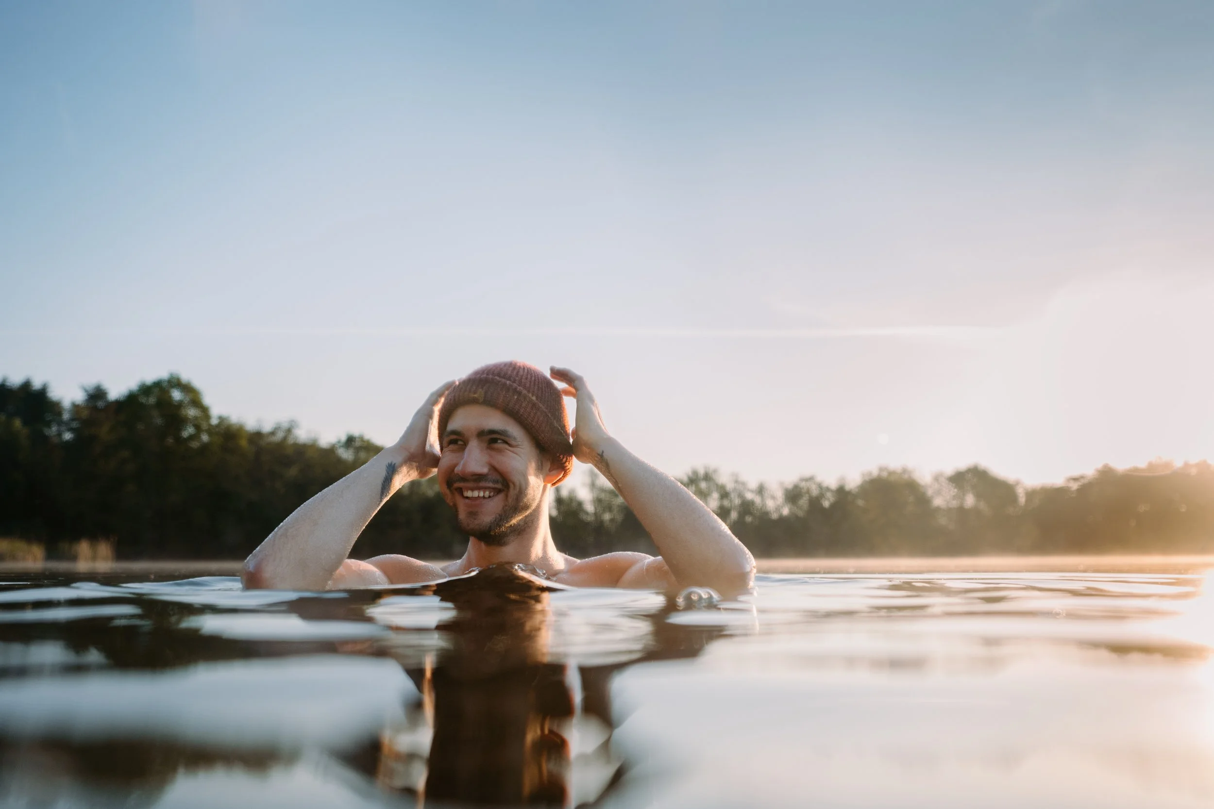 A man smiling in a body of water during sunset, wearing a knit hat, with a natural landscape in the background.