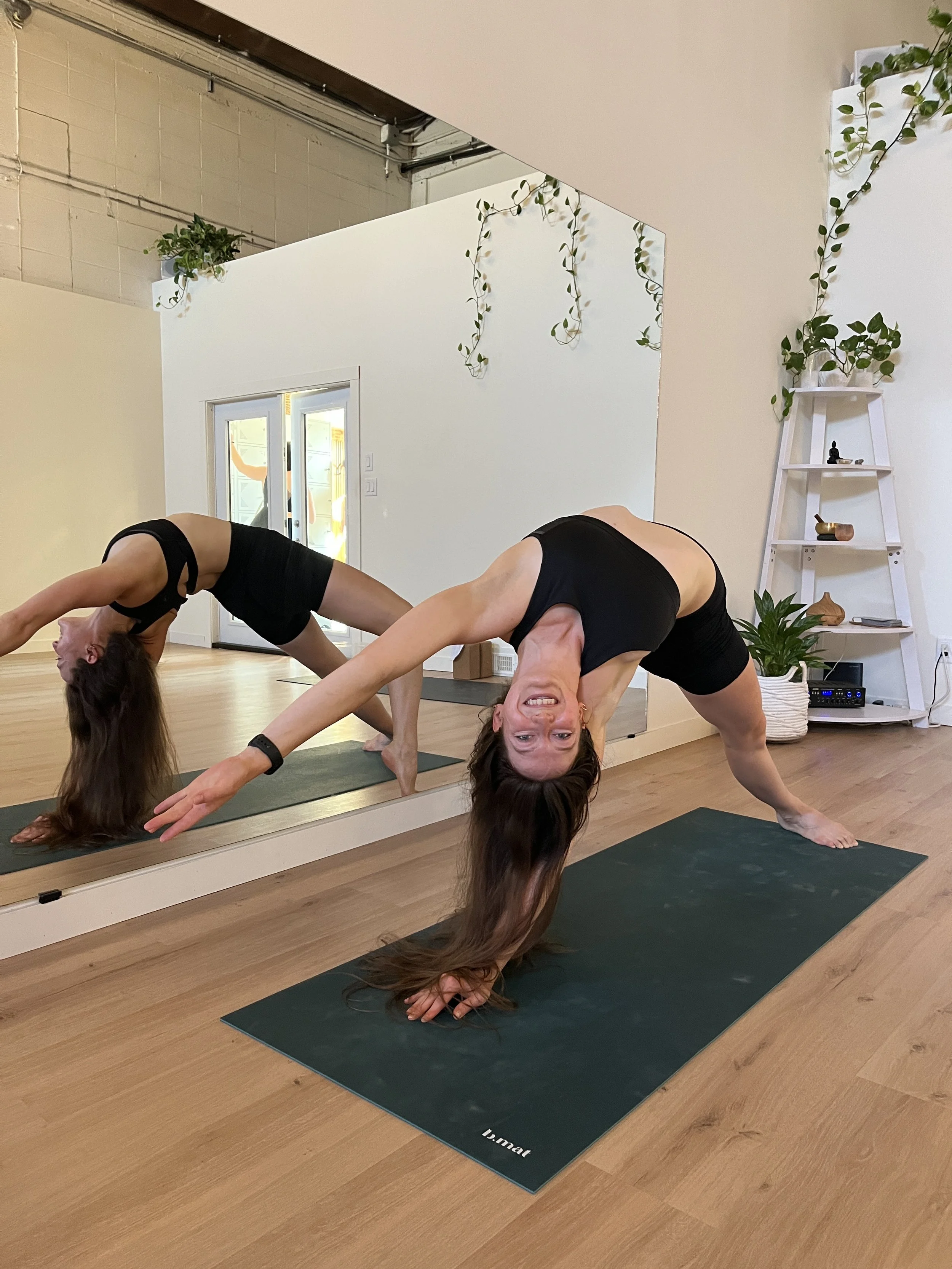 Two women practicing yoga in a studio with fitness mats, a large mirror, and a white wall decorated with plants.