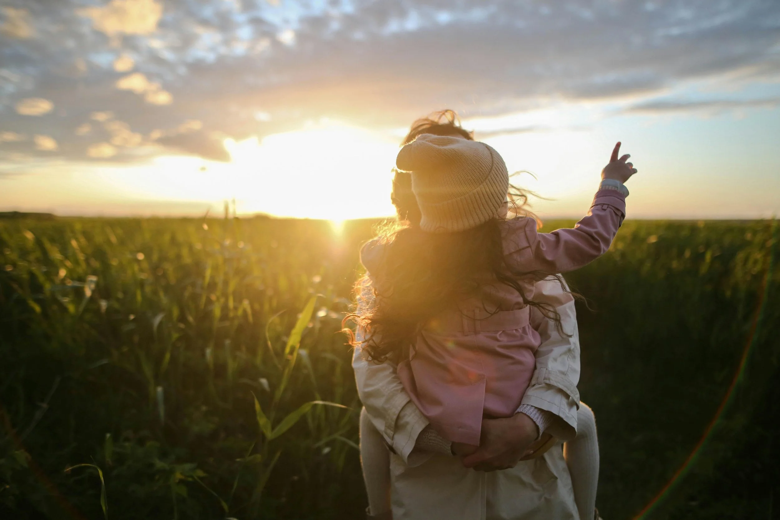 Person carrying a child in a field during sunset, with the child's arm raised and pointing.