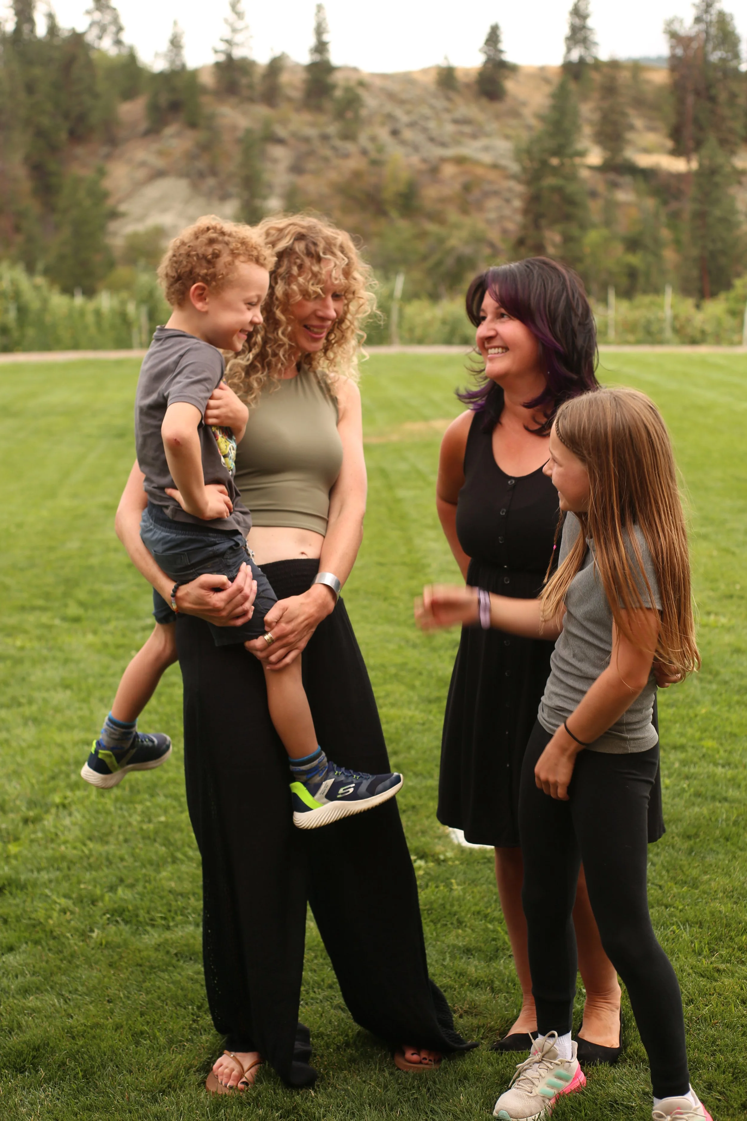 Four people, two women and a girl, laughing and talking outdoors on a grassy field with trees and hills in the background.