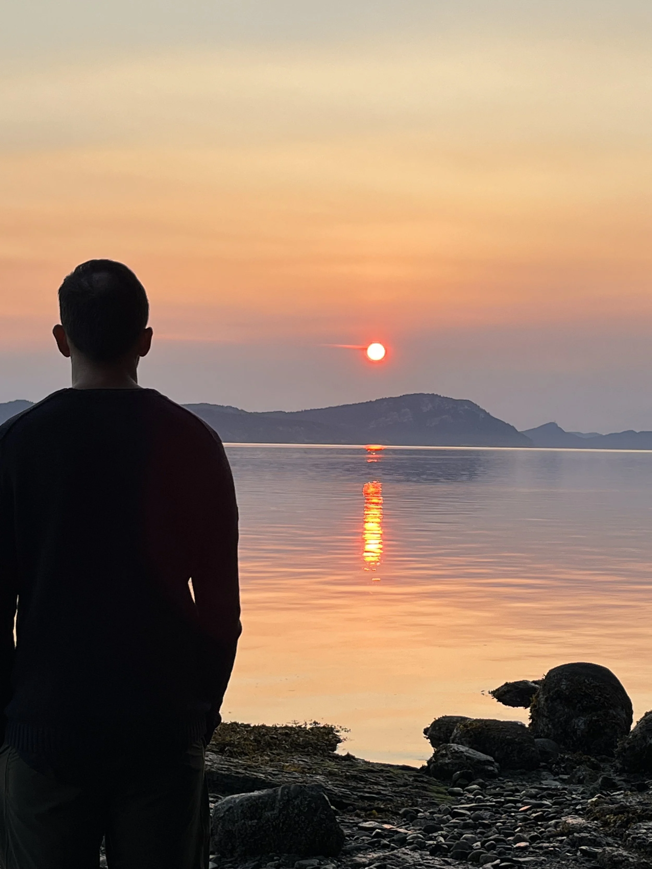 Silhouette of a man standing on a rocky shoreline, watching a sunset over a calm lake with mountains in the distance.