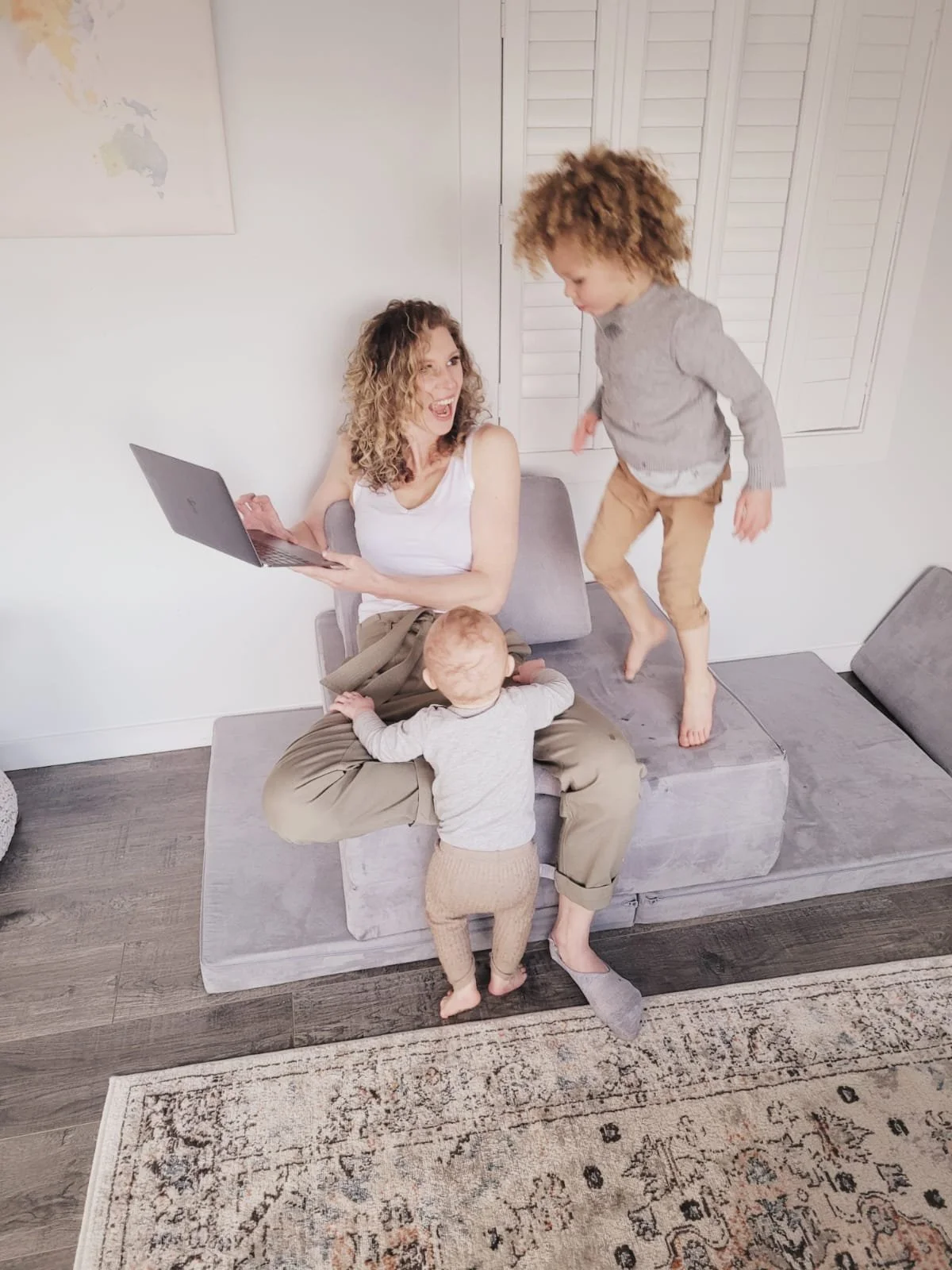 A woman sitting on a couch holds a laptop and looks joyfully at two children, one standing and one jumping on her lap, in a living room with a world map on the wall.