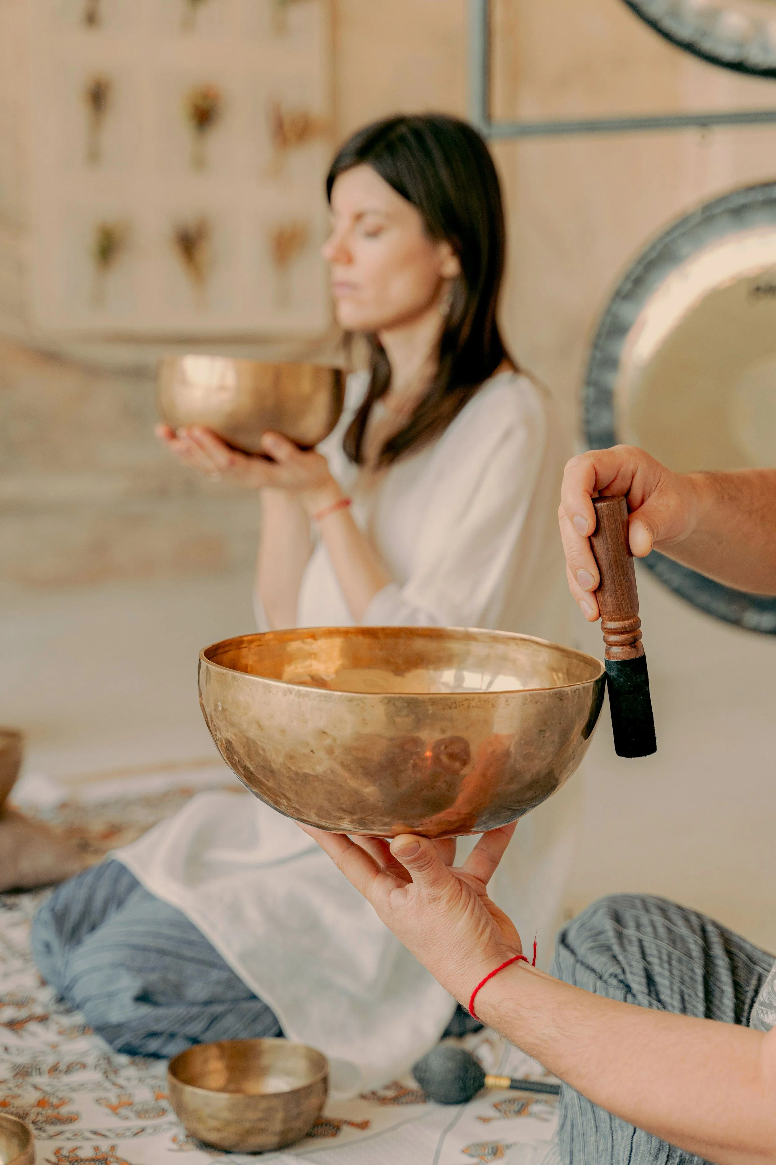 Two people holding Tibetan singing bowls in a space decorated with small potpourri containers. One person is sitting with eyes closed, the other is holding a mallet.