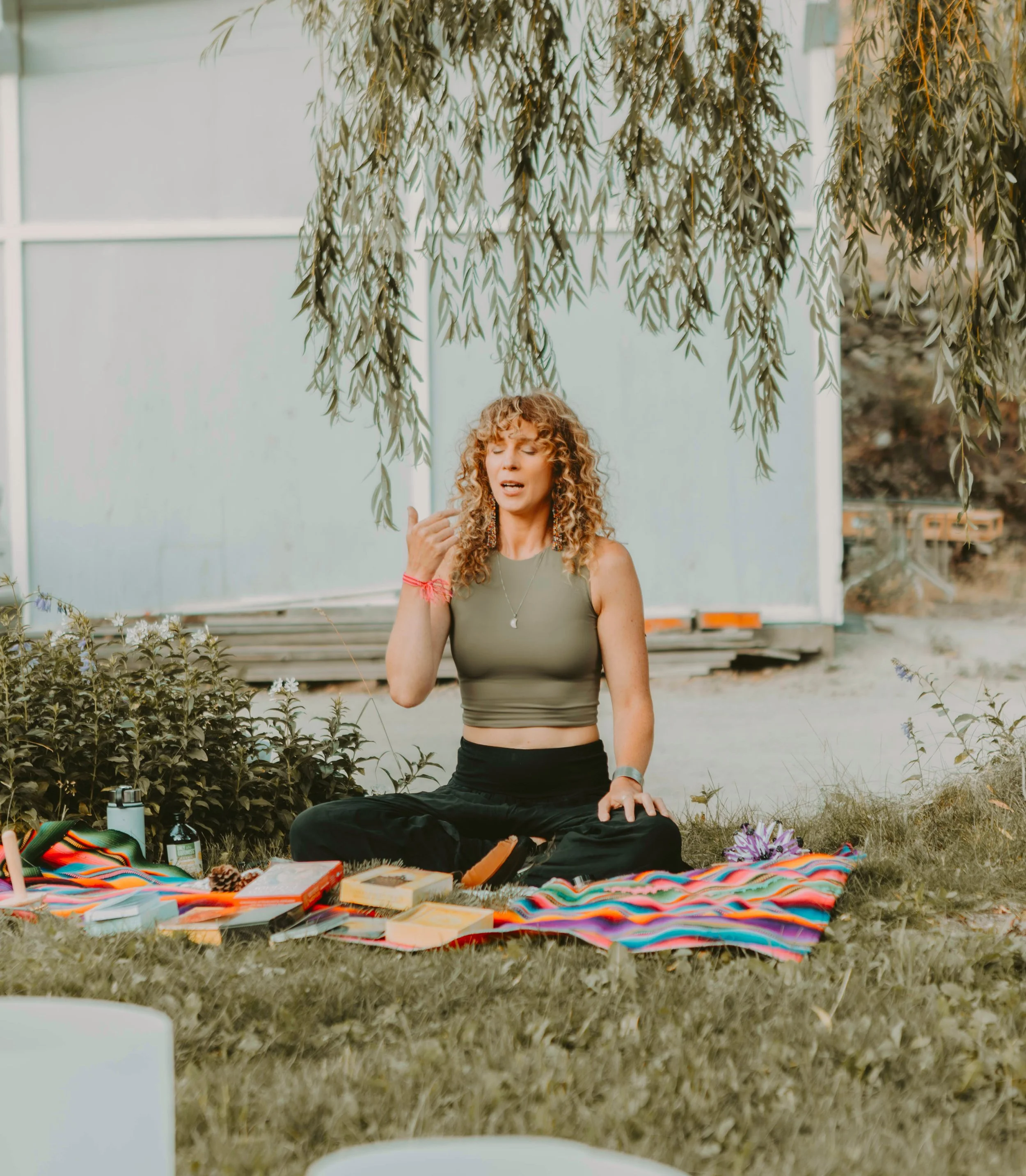 A woman with curly hair practicing yoga outdoors, seated cross-legged on a colorful striped blanket under a tree, with her eyes closed and one hand near her face, surrounded by books, water bottles, and small objects.