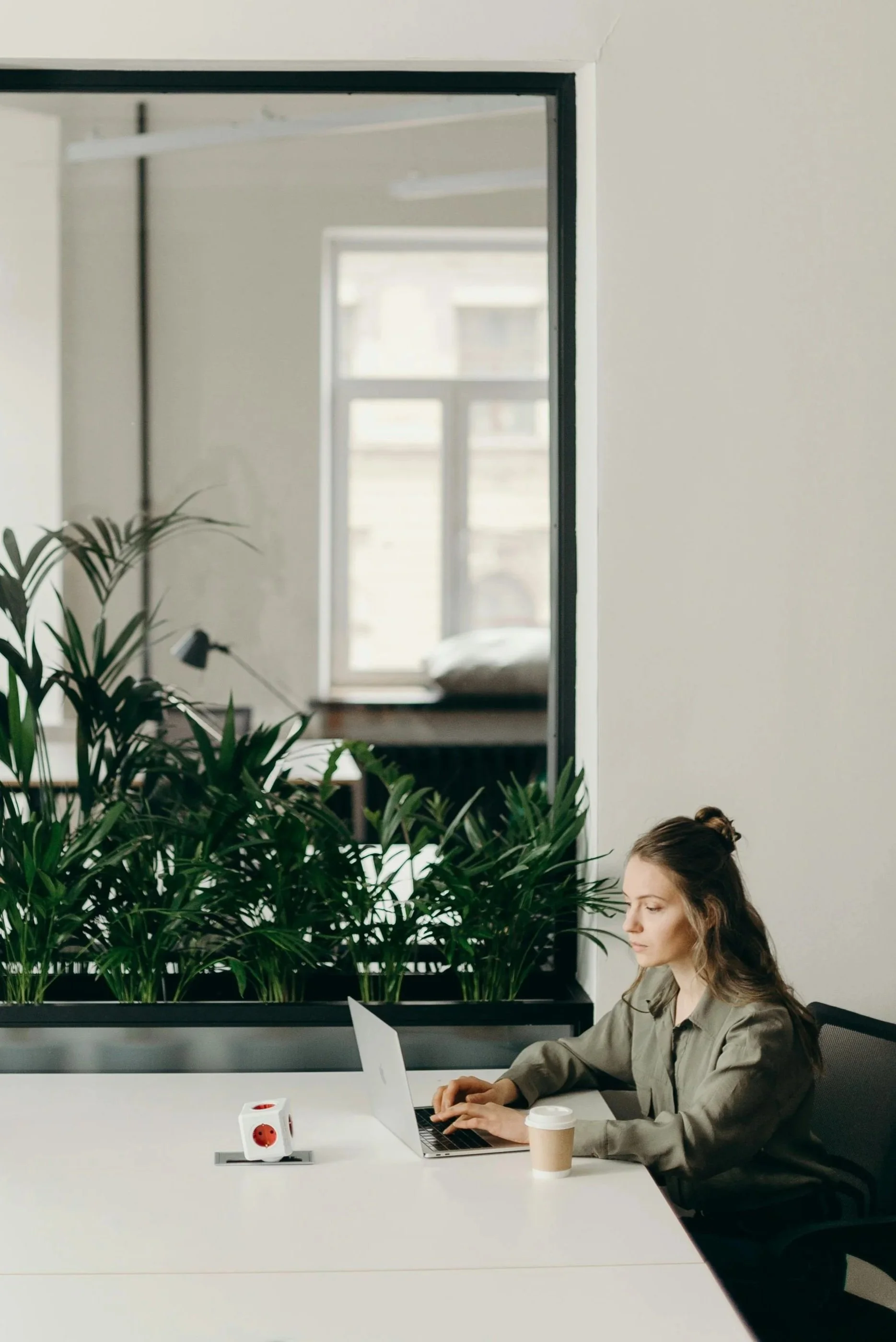 A woman working on a laptop at a white desk with a coffee cup, a decorative dice, and a row of green plants in a modern office space.