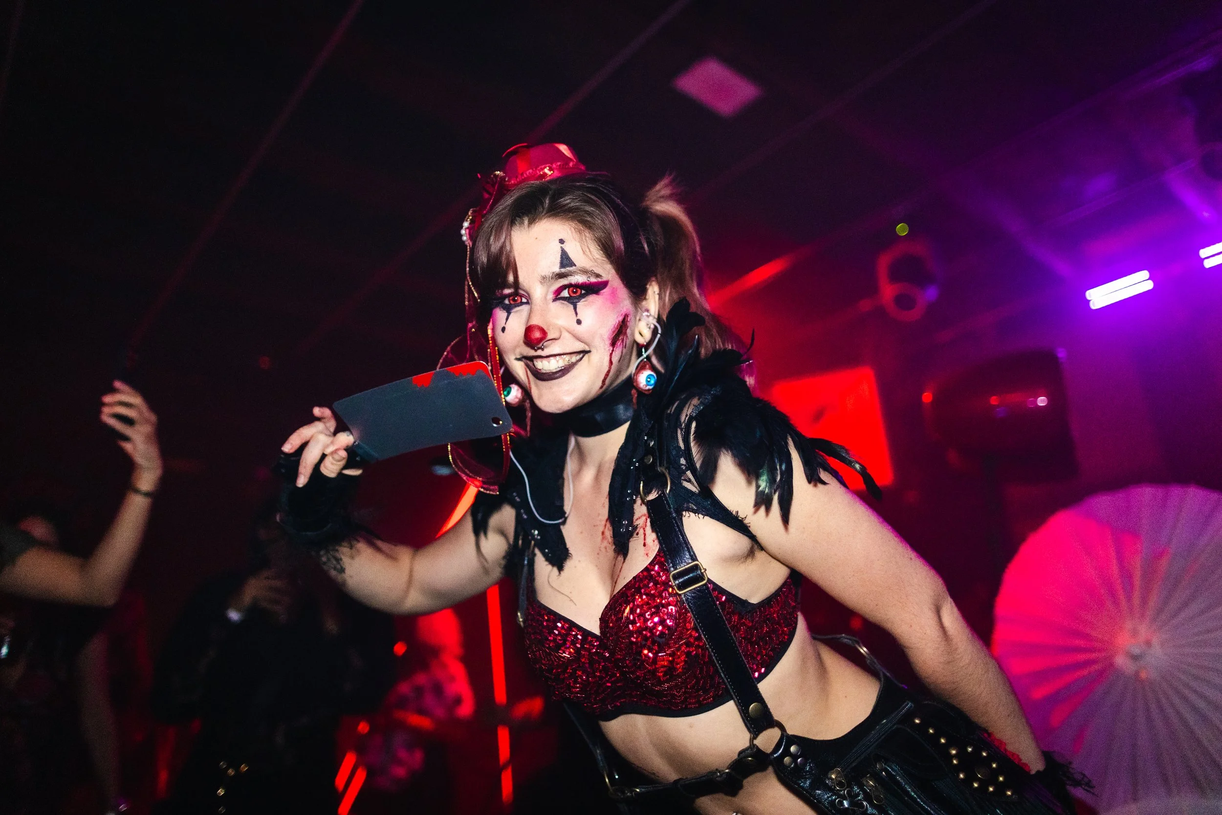 Woman dressed as a sinister clown with makeup, red nose, and wig at a party with red and purple lighting.