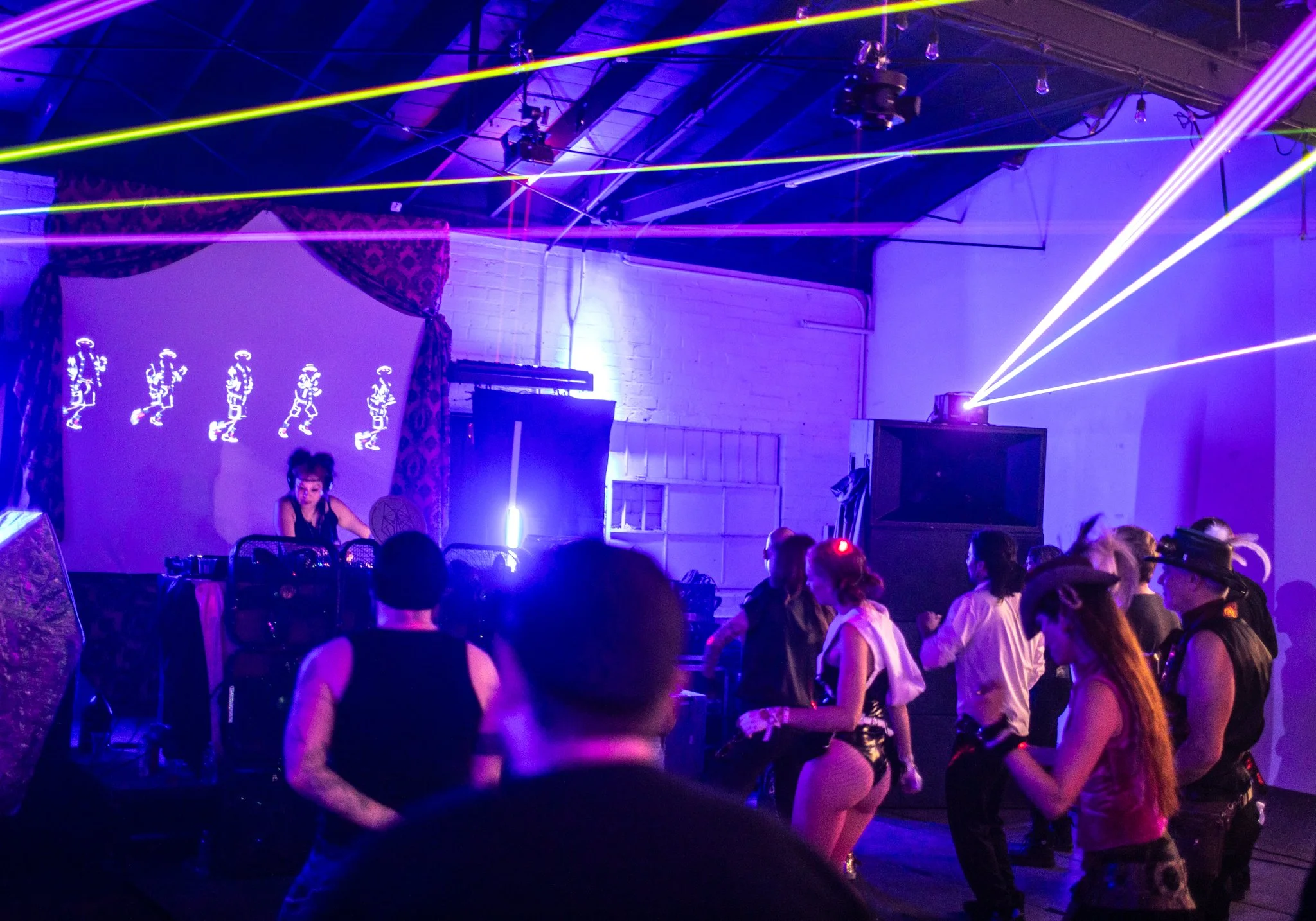 People dancing at a neon-lit electronic music event with colorful laser lights and a DJ performing on stage.