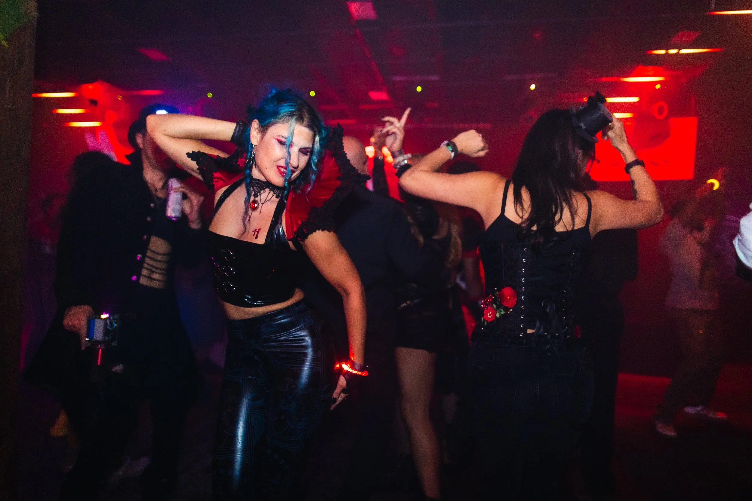 People dancing at a nightclub, with vibrant red and purple lighting, dressed in dark, stylish clothes, some wearing accessories like top hats and floral embellishments.