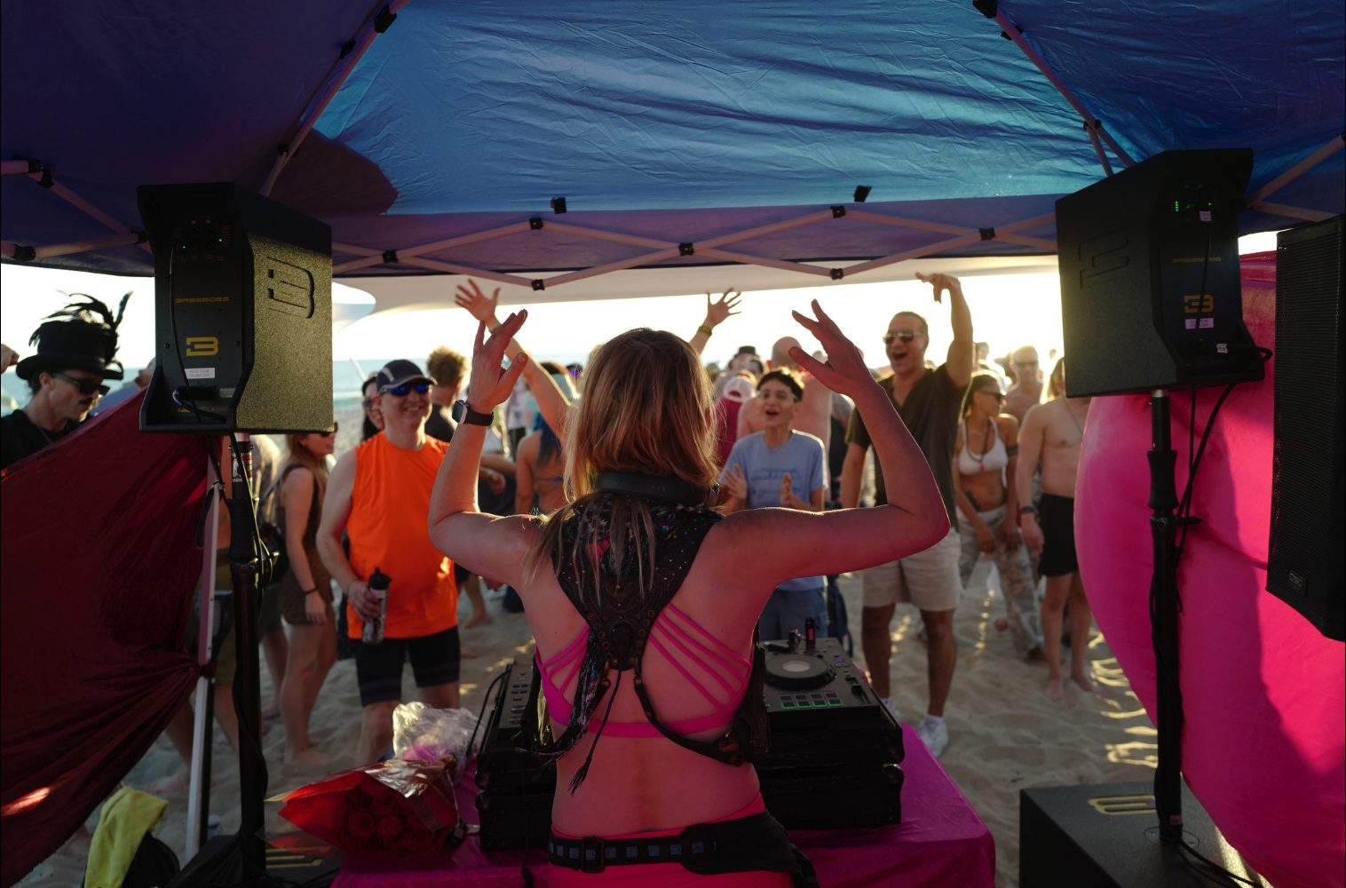A woman DJ performing at a beach party with a crowd dancing; the scene is illuminated by sunset light.