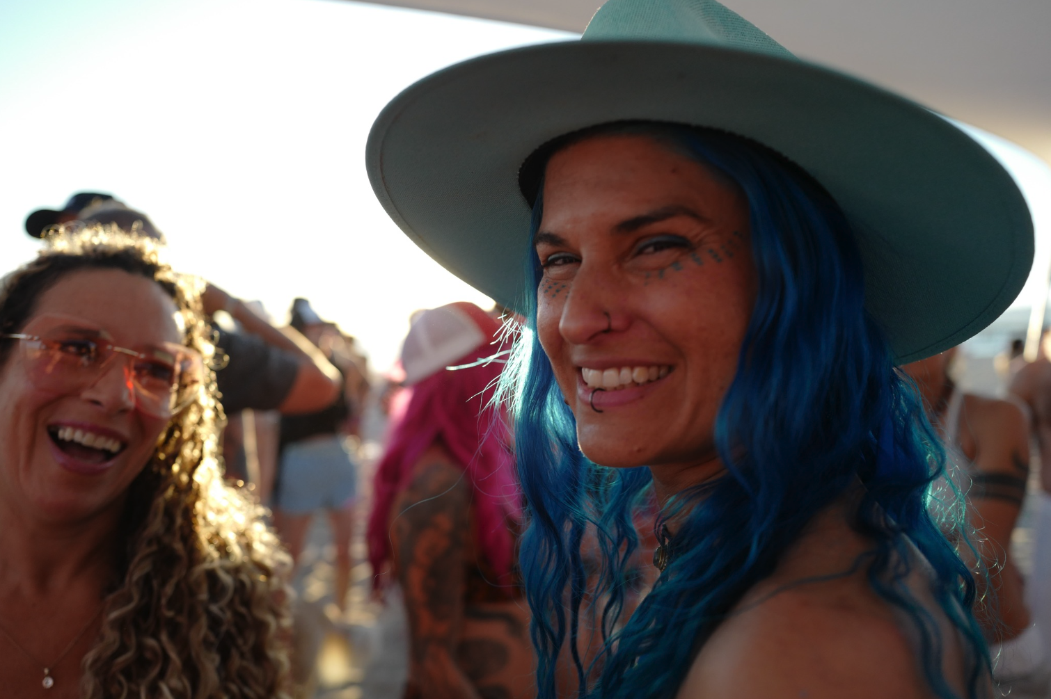 Two women smiling at an outdoor event, one with blue hair and a large hat, the other with curly hair and glasses, in a crowd during sunset.