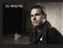 Portrait of DJ Wolfe with a serious expression, wearing a dark jacket and scarf, in an industrial-style setting.