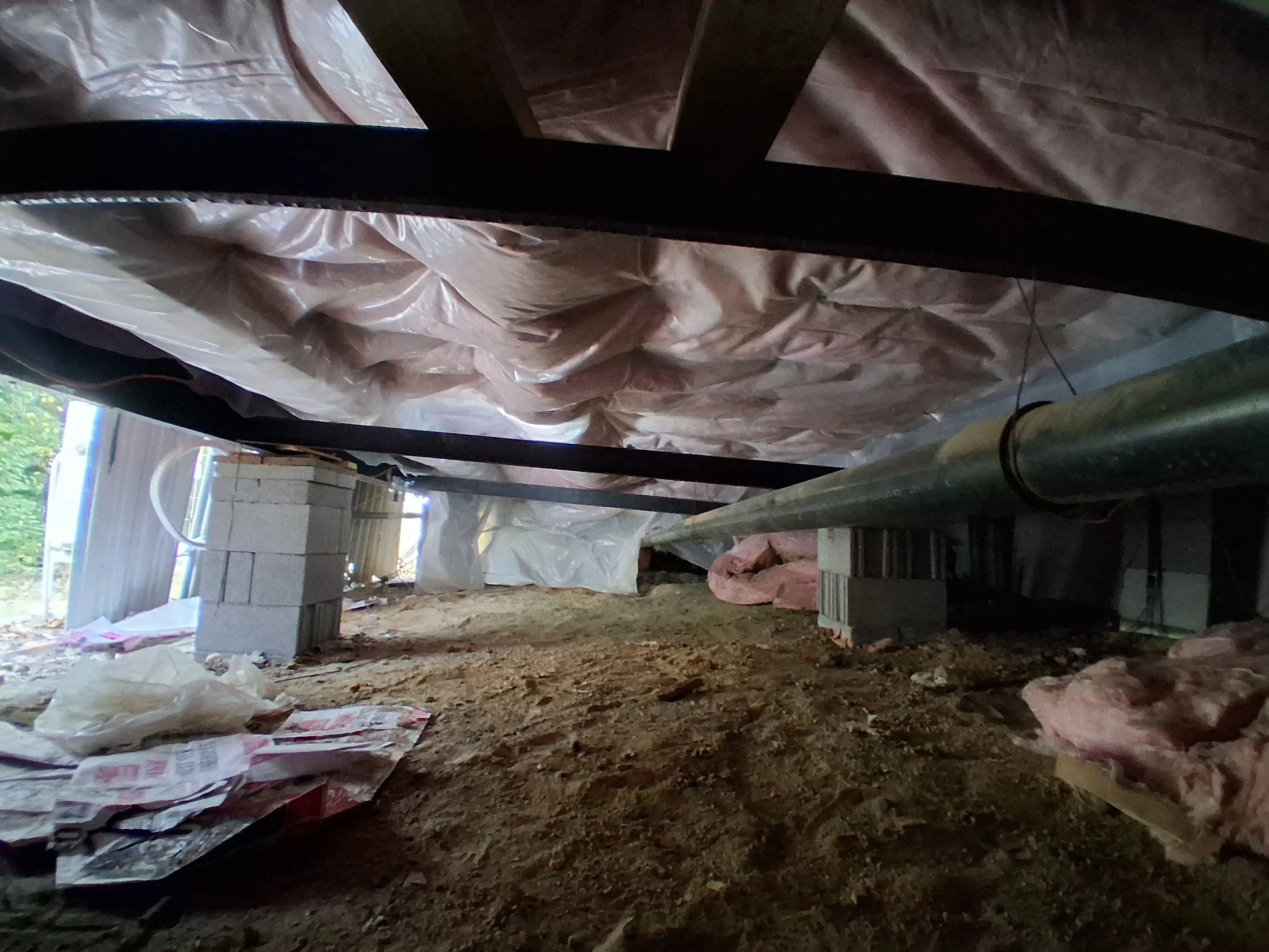 View of a construction site from within a crawl space showing insulation, a metal pipe, cinder blocks, and exposed soil.