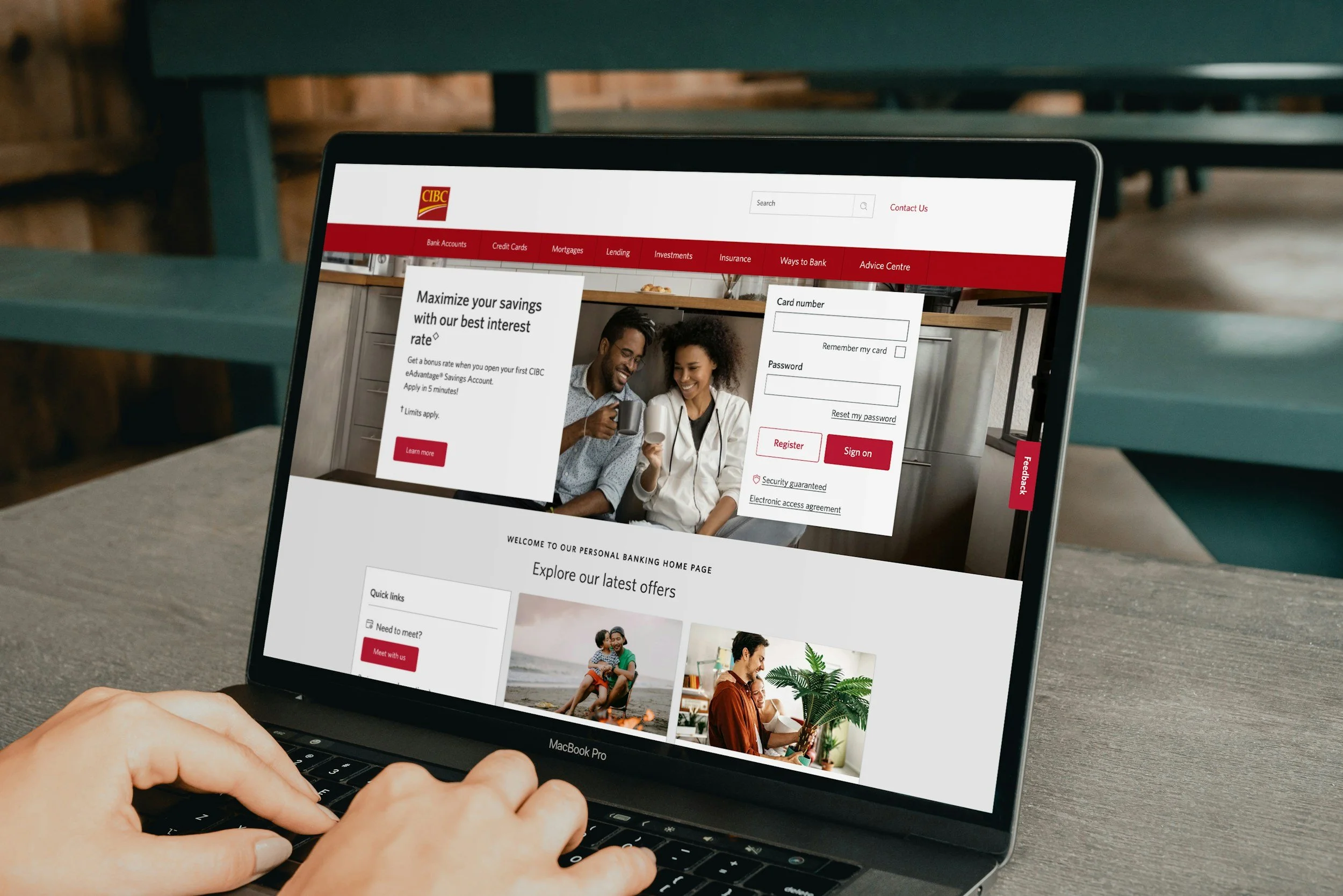 Laptop displaying the CIBC online banking login page, with options to log in or register, featuring a photo of two people smiling and coffee mugs, on a wooden table with a blurred background.