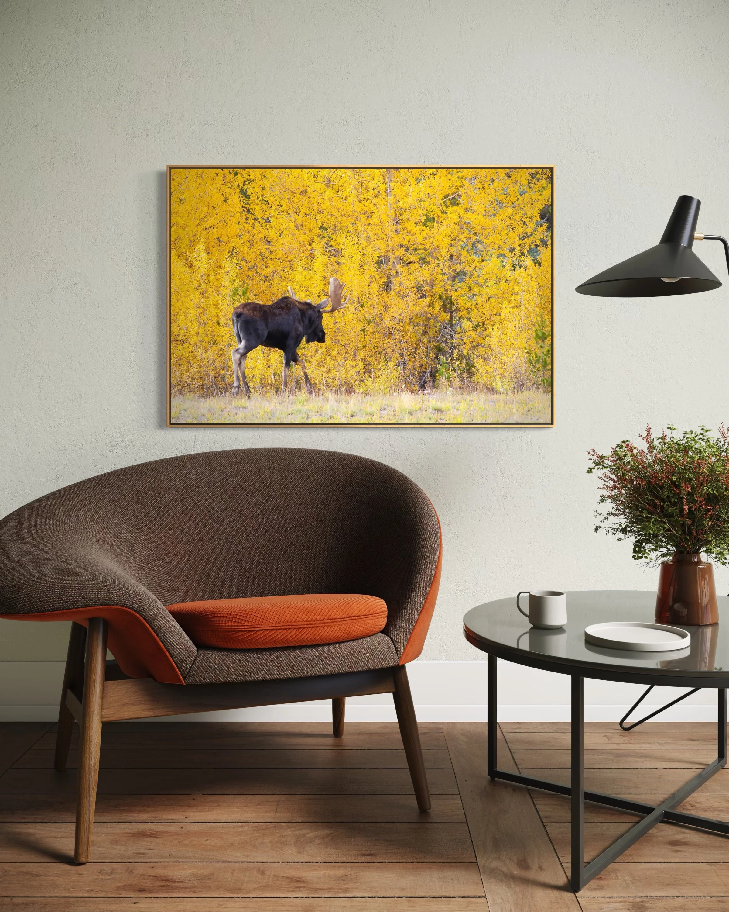 Living room with a modern armchair, a side table with a drink and a plate, a lamp, and a wall art depicting a moose in a forest during fall.