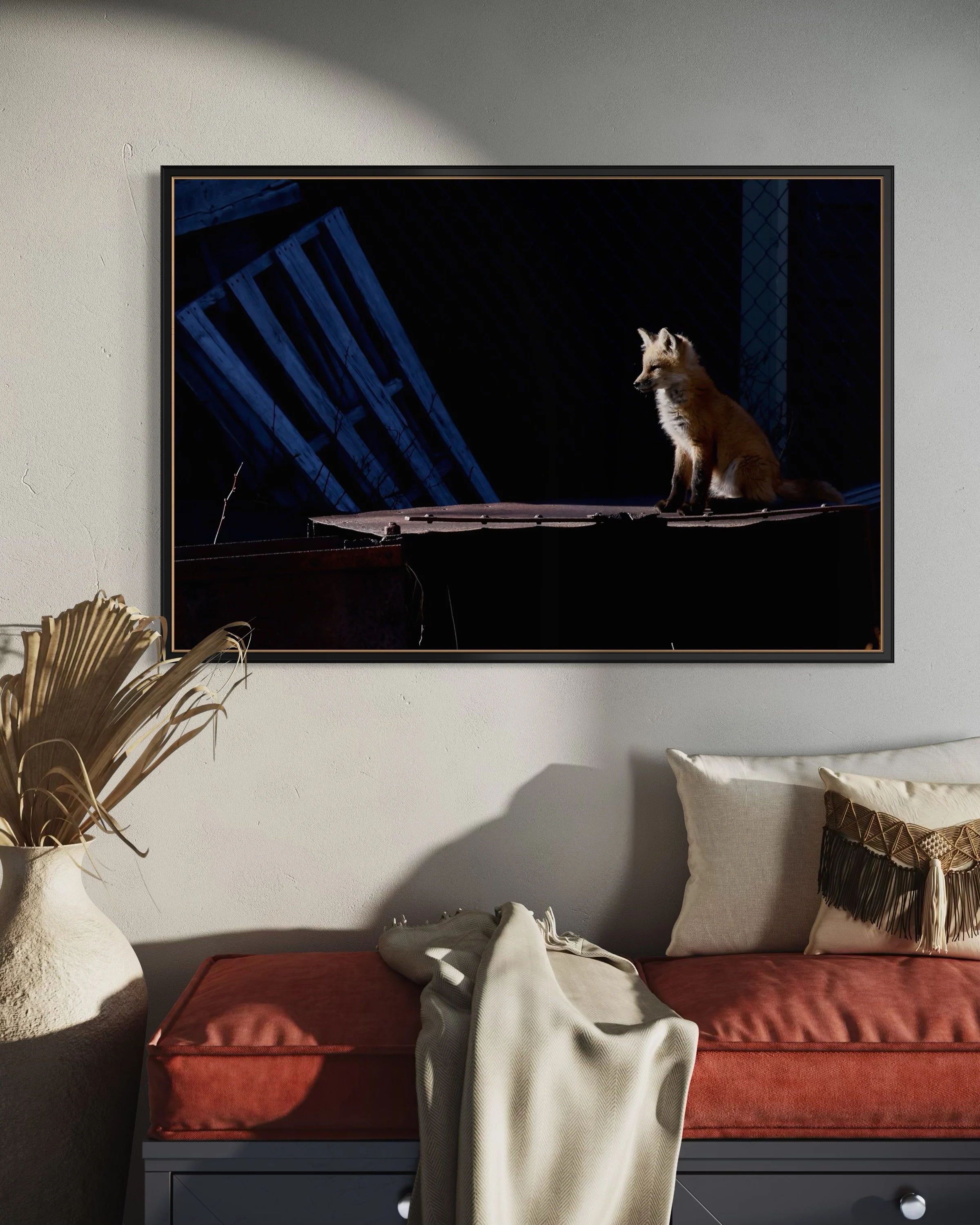 Photo of a sofa with pillows in a living room, a framed picture of a fox on the wall, and a large vase with dried plants beside it.