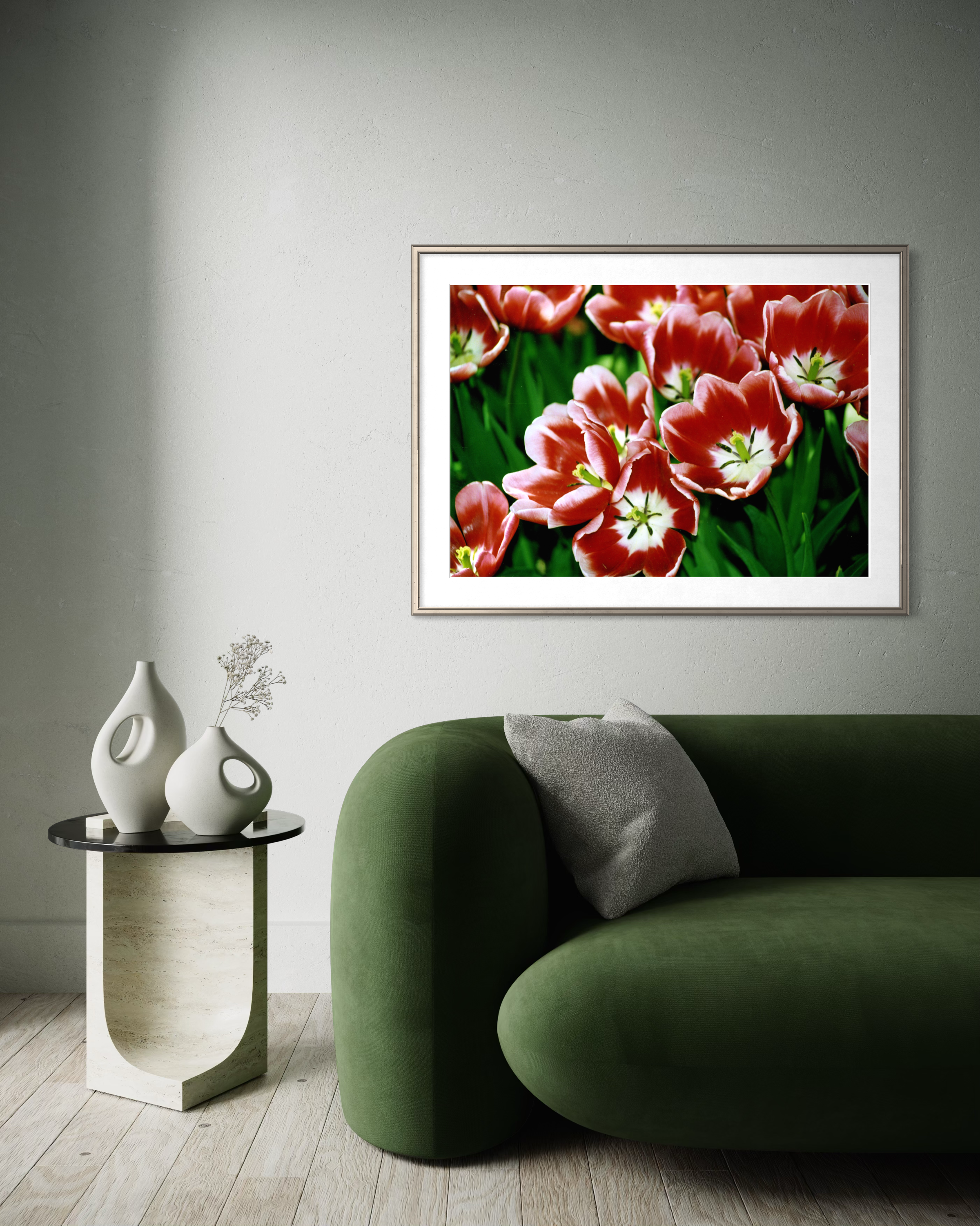 Living room corner with a green upholstered seating and a white side table holding two white vases with decorative branches, wall art of pink tulips with green leaves, and wooden flooring.