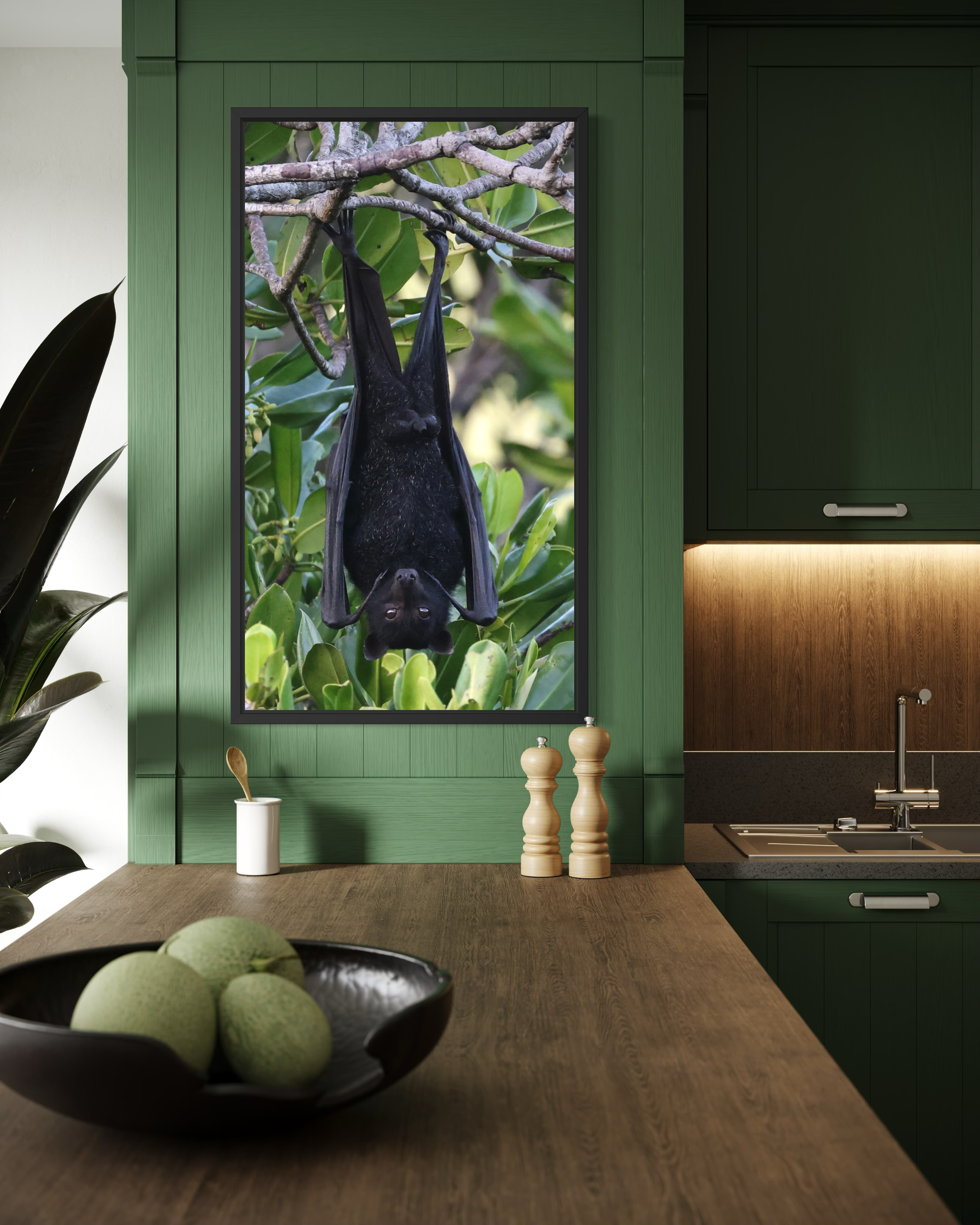 A modern kitchen with a wooden dining table, green cabinets, decorative vases, and a framed picture of a black bat hanging upside down on a leafy tree.