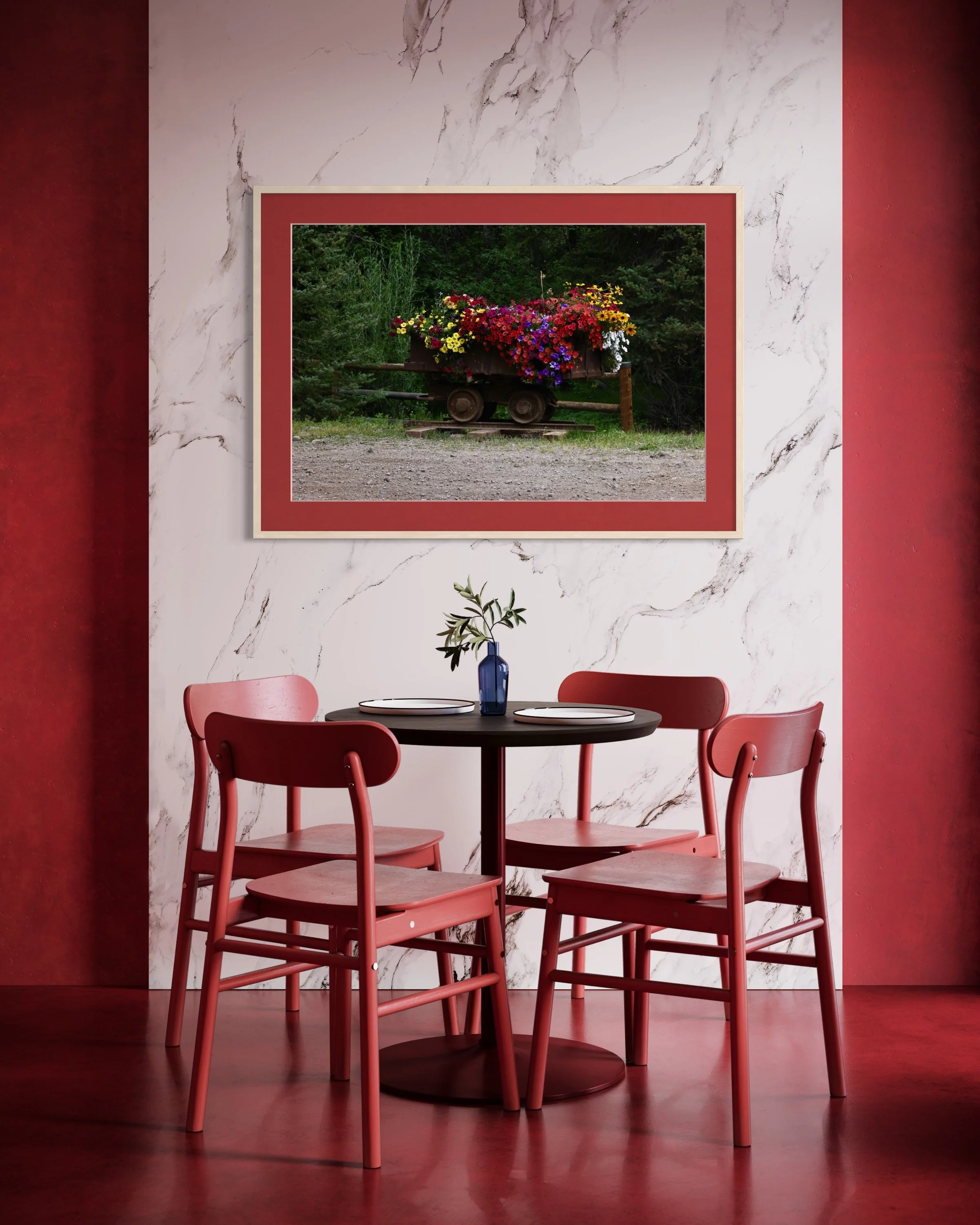 A round black table with four pink chairs in a room with a marble wall and red side walls. On the wall, there is a framed painting of a wagon filled with colorful flowers. A small plant in a blue vase is on the table.