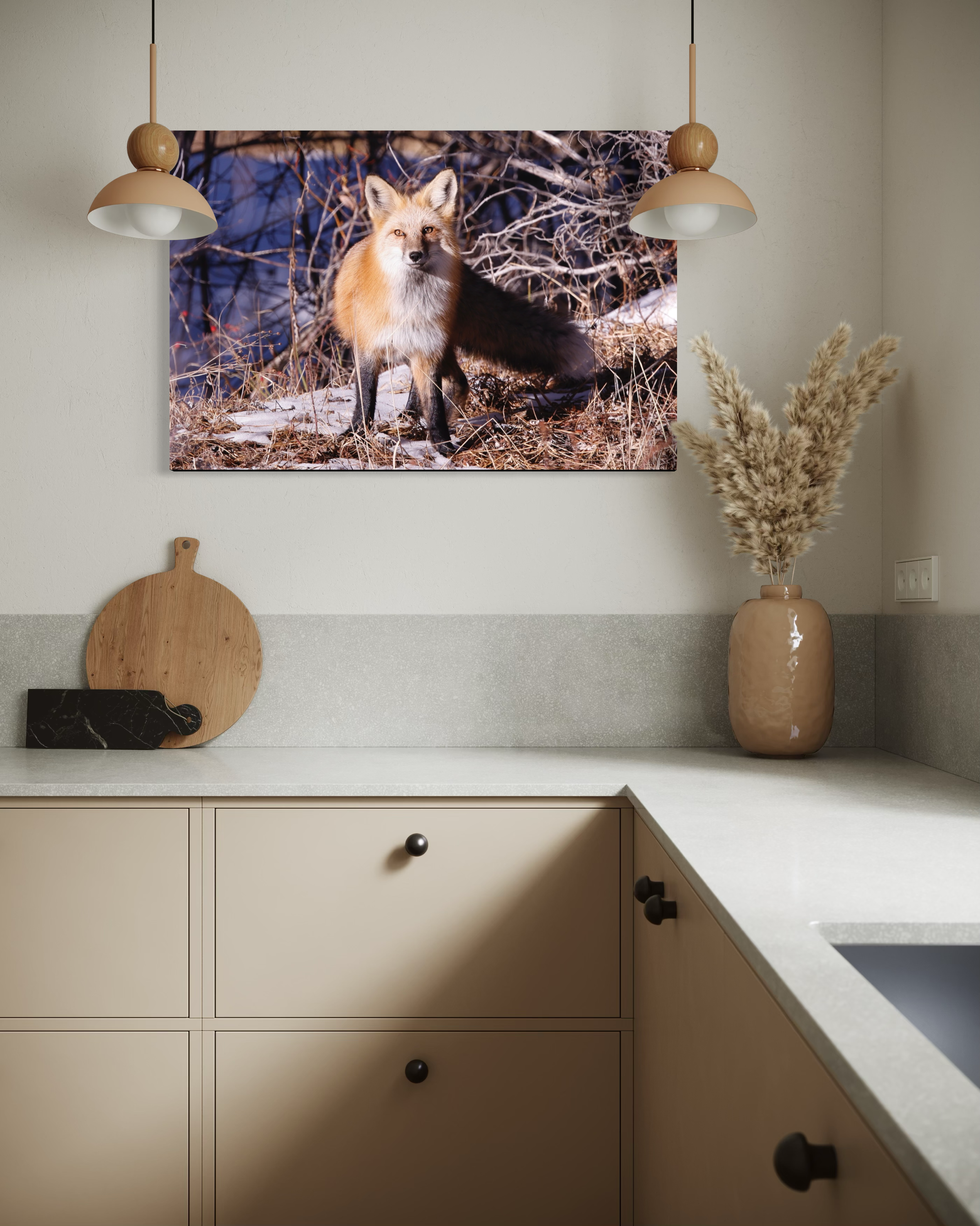 A kitchen with beige cabinets and a white countertop, topped with a large beige vase holding tall dried pampas grass. There is a wall with a power outlet in the corner and a small black object on the countertop in the bottom left. A large photo of a 