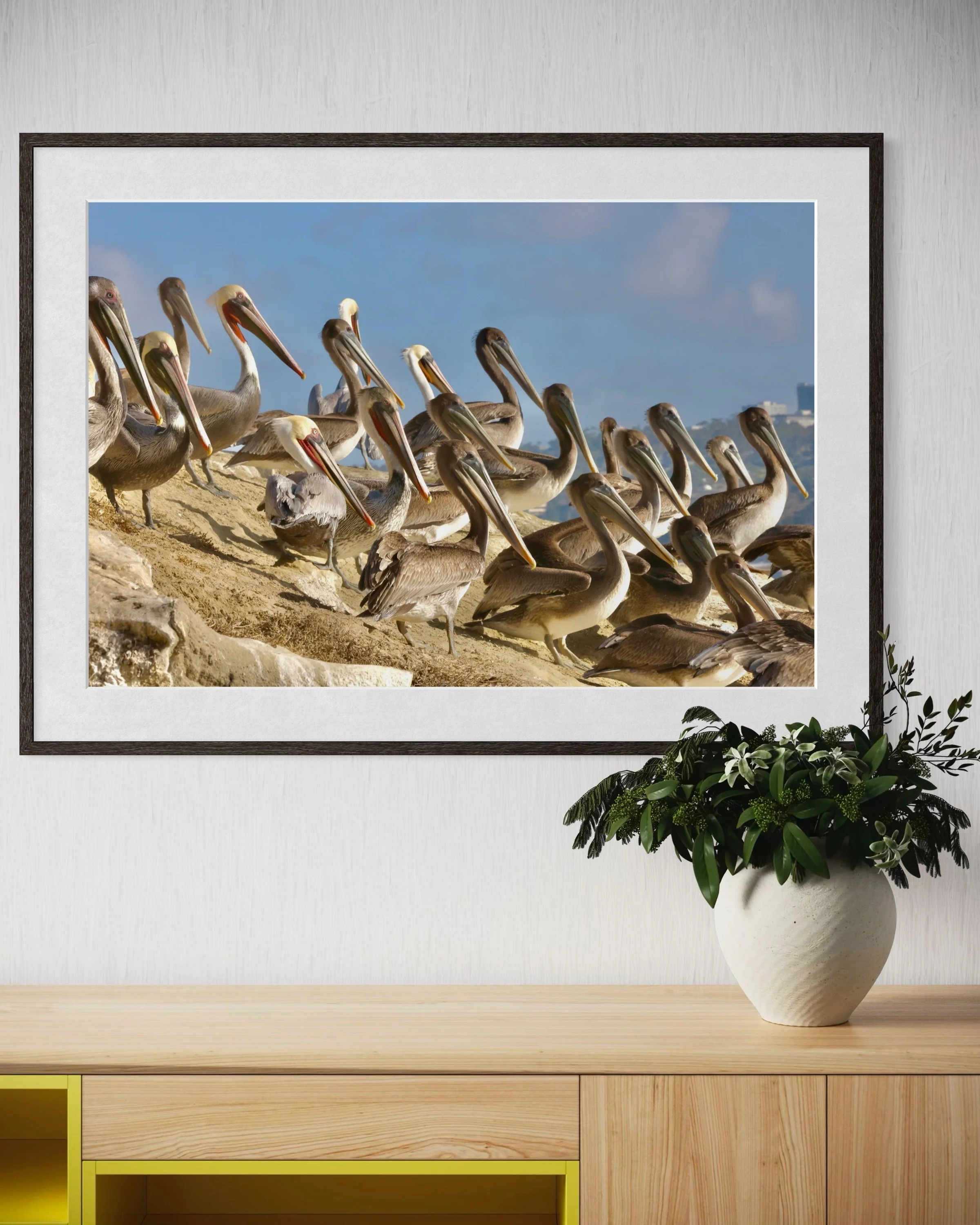 A room with a framed picture of pelicans on the wall above a light wooden cabinet. There is a white vase with green and dark leaves on the right side of the cabinet.