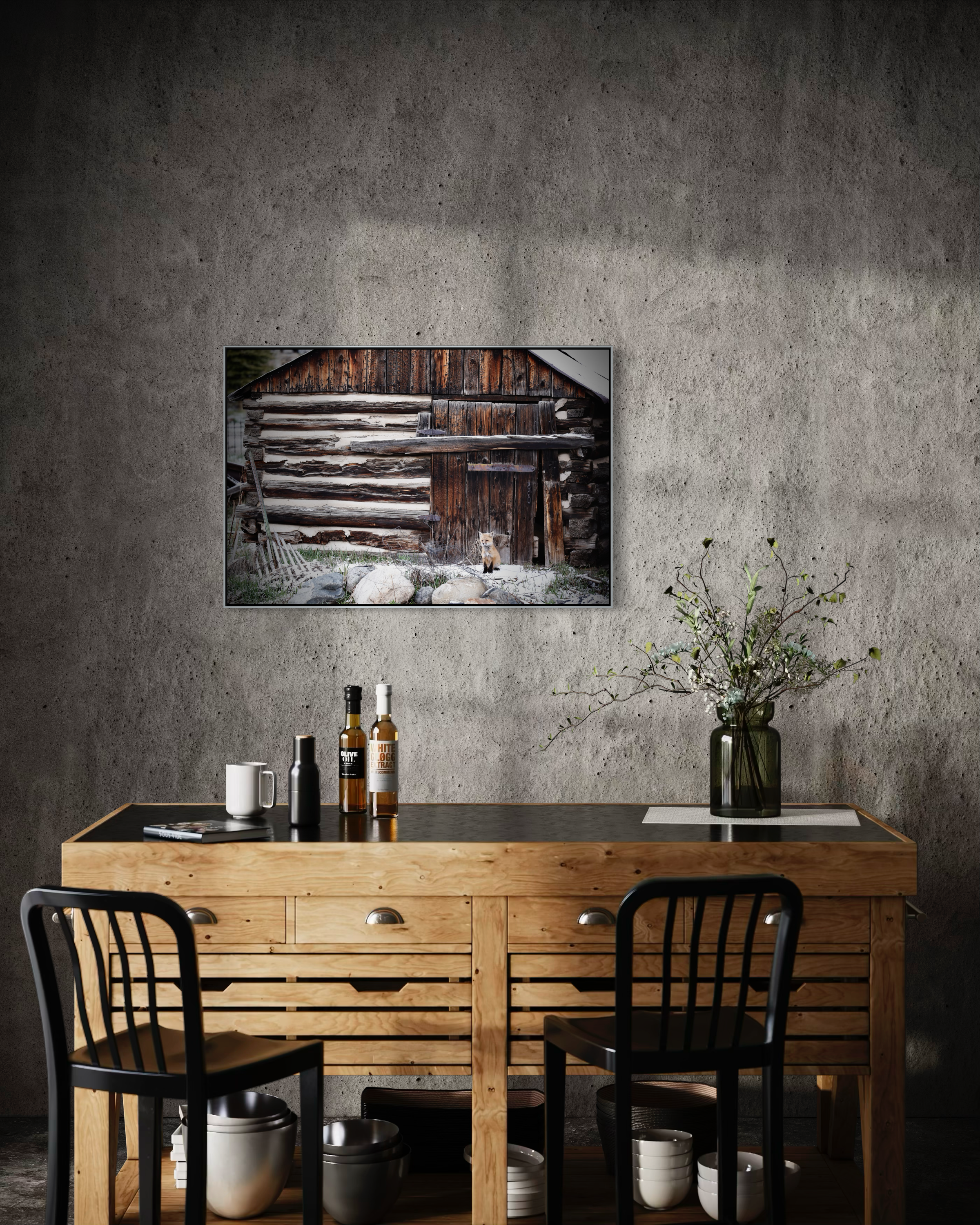 A wooden dining table with four black chairs, a black table surface, and items including bottles of olive oil and white sugar, a white mug, a black bottle, a stack of books, and a glass vase with green branches on a placemat. A large framed photo of 