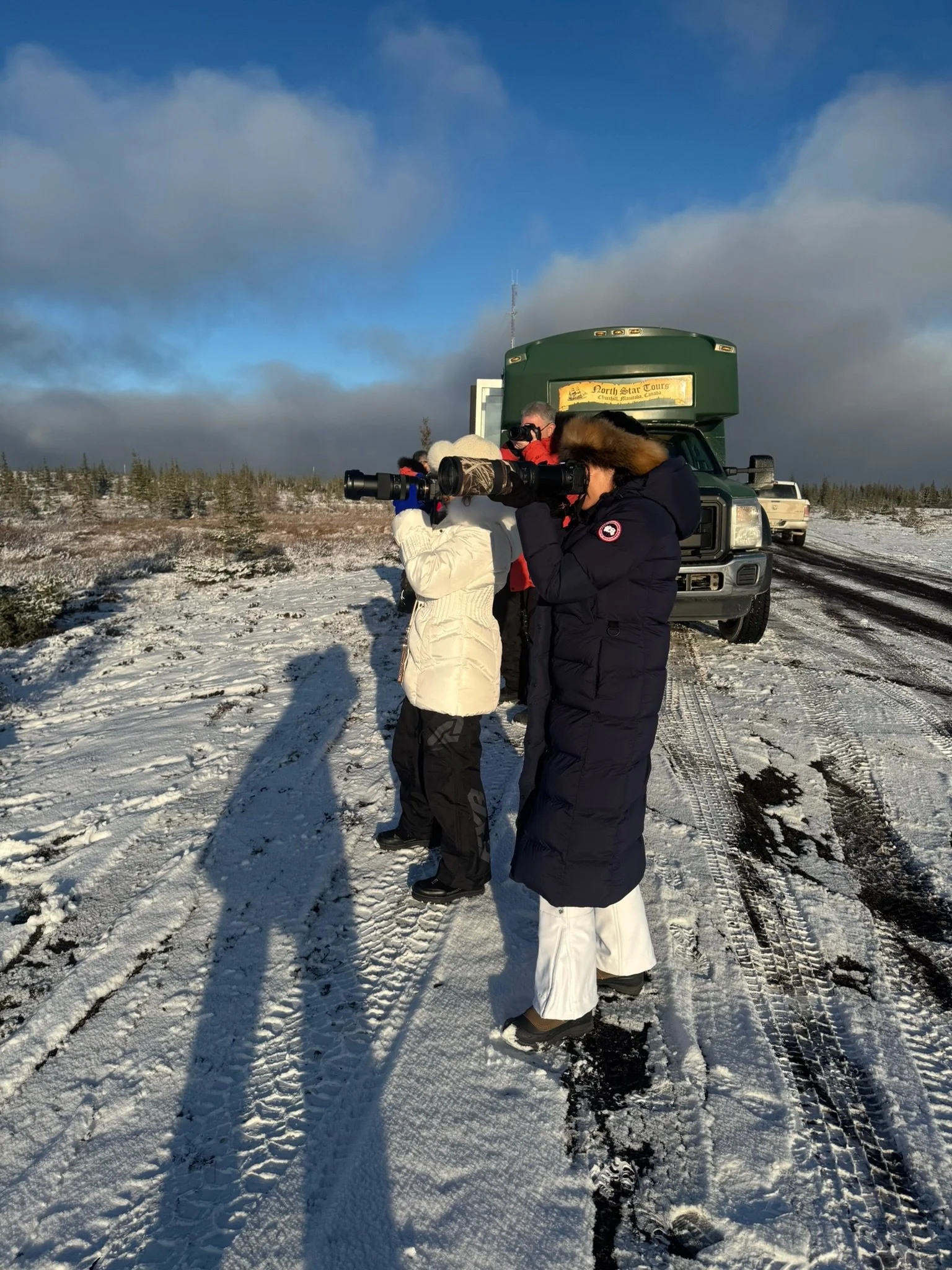 People in winter clothing using binoculars and camera, snow-covered landscape with trees, parked trucks, cloudy sky.