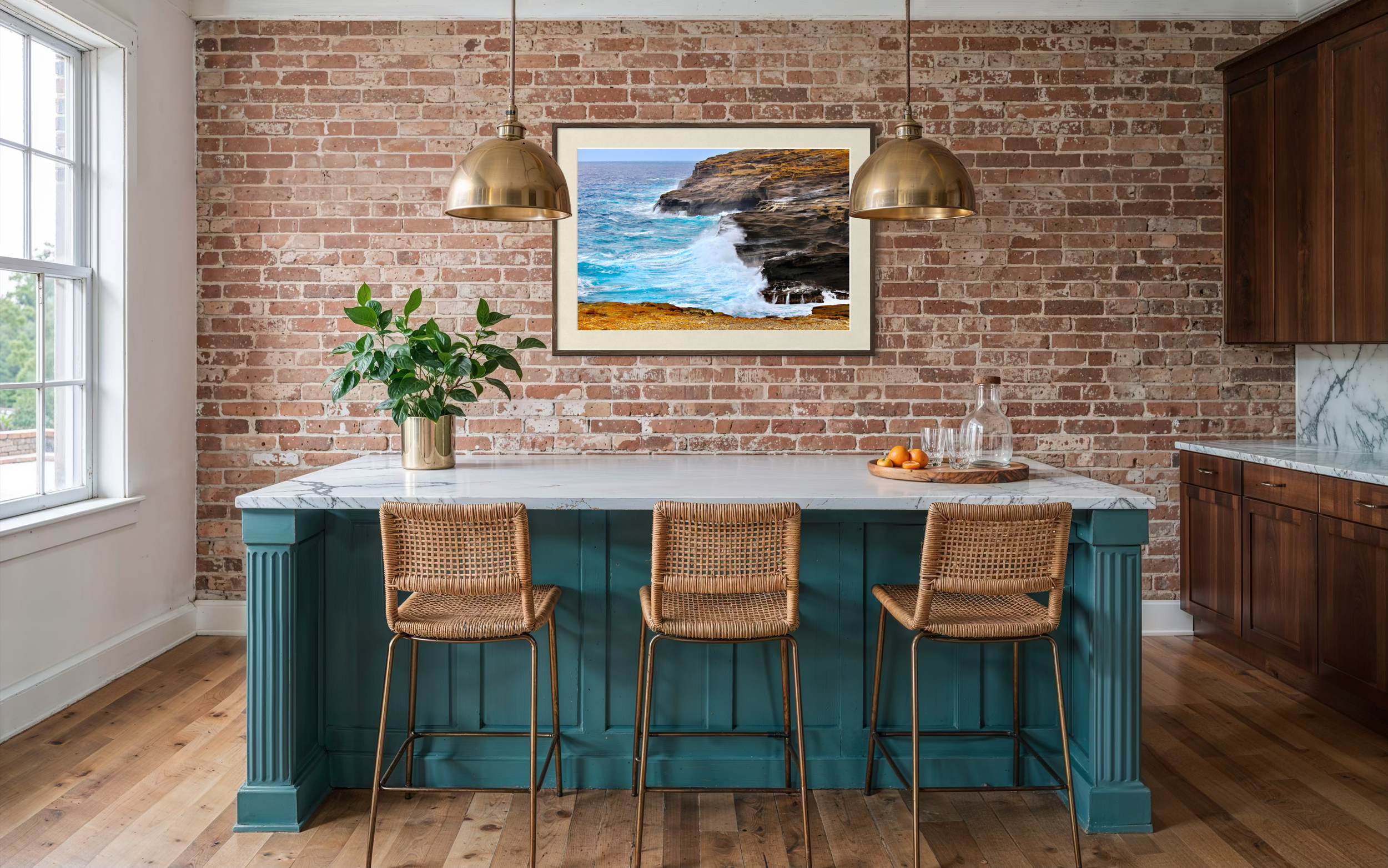 Interior of a kitchen with a brick wall, a framed landscape photo of the ocean and rocks, a teal kitchen island, three rattan chairs, a green potted plant, and wooden cabinets.