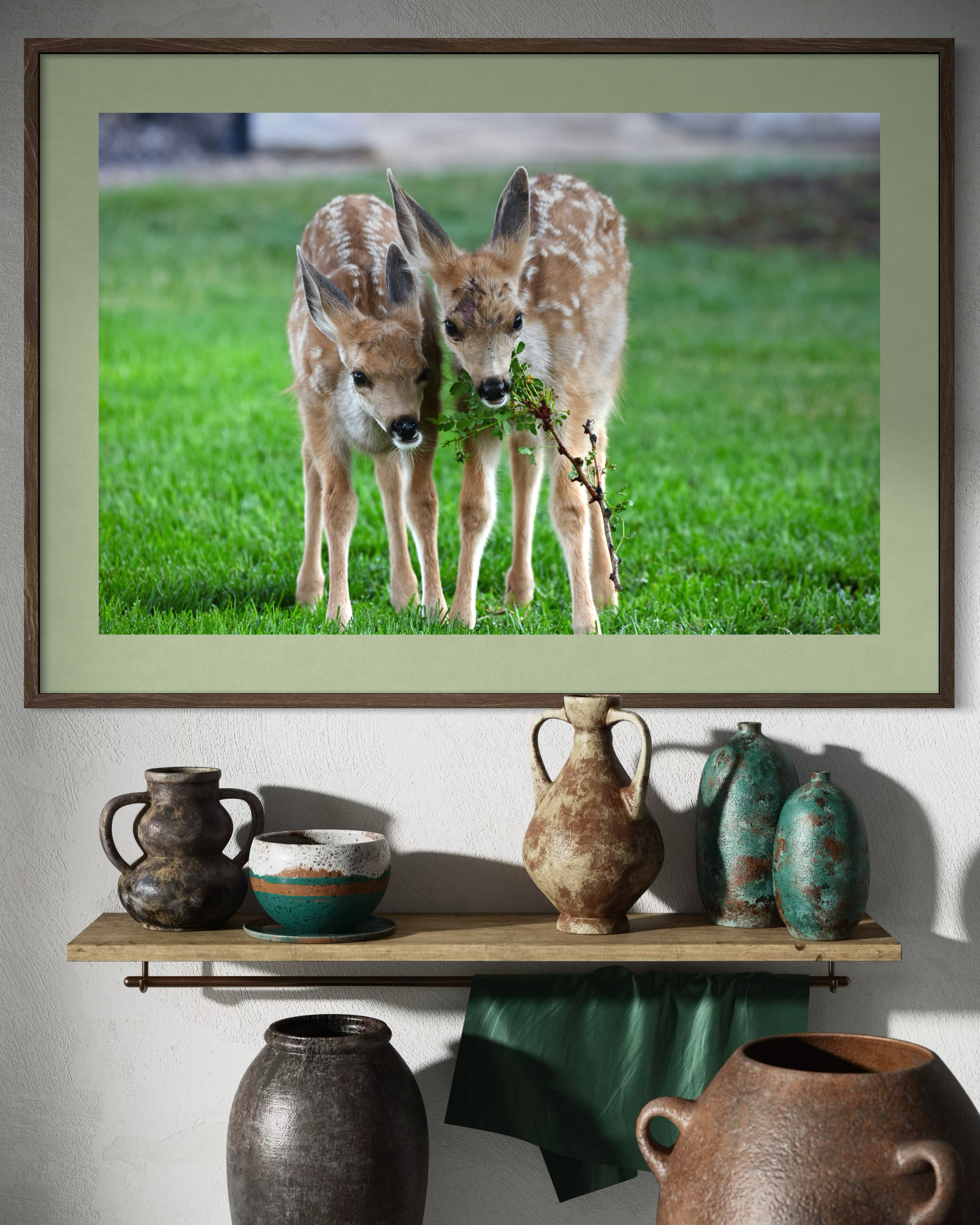 A framed photograph of two young deer with light brown fur and white spots, standing on green grass, one holding a small branch with leaves in its mouth.