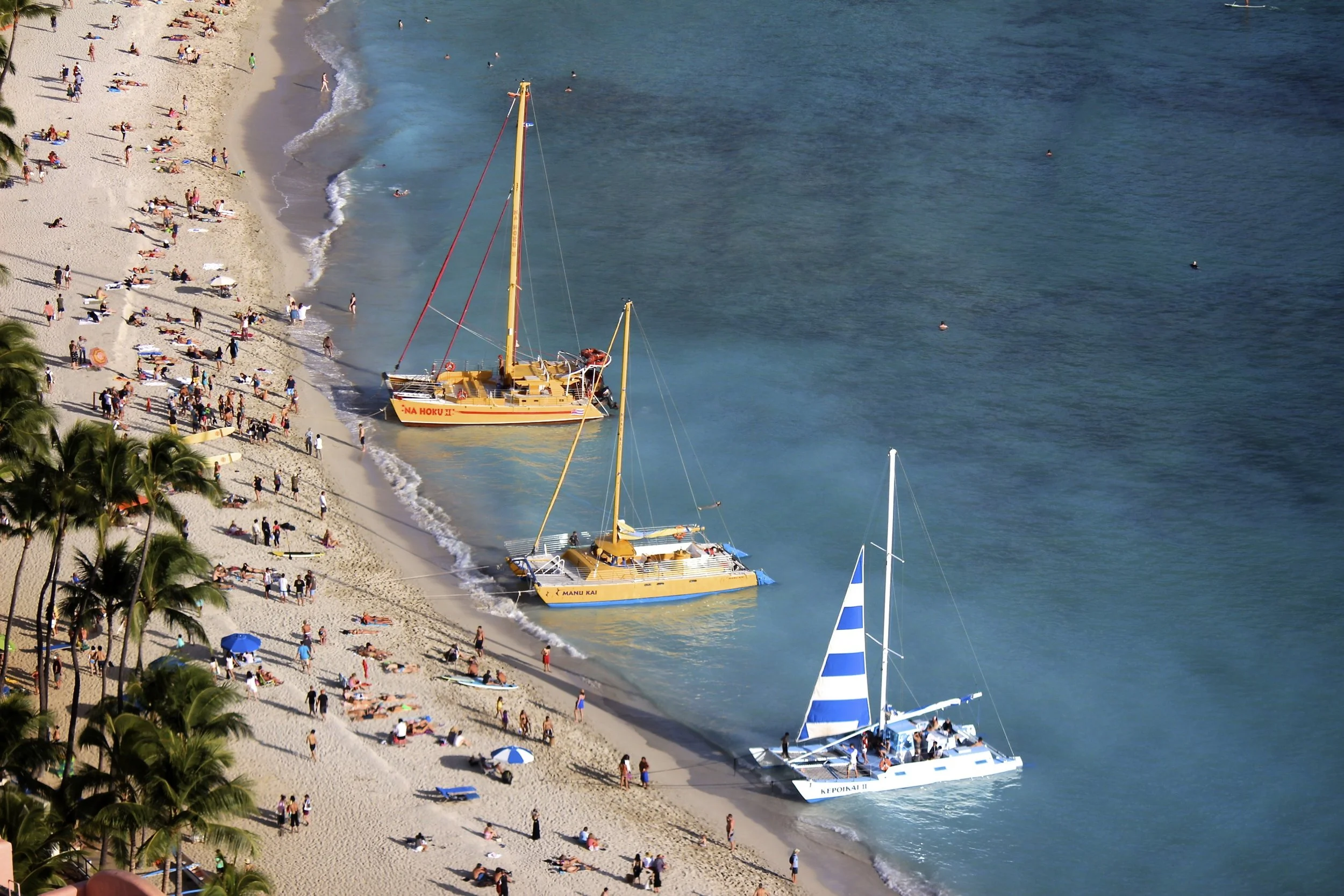 A busy beach scene with numerous sunbathers and visitors on the sandy shoreline, colorful umbrellas, palm trees along the coast, and three boats docked on the water representing a lively seaside area.