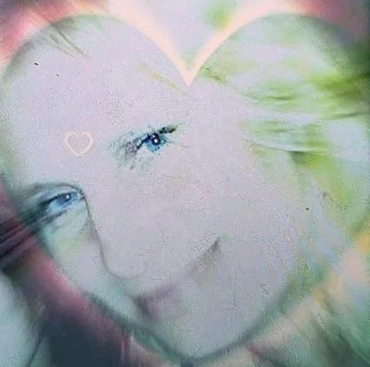 A woman with light-colored eyes and blonde hair, with a small heart shape drawn on her forehead, surrounded by a soft, colorful, and blurred background.