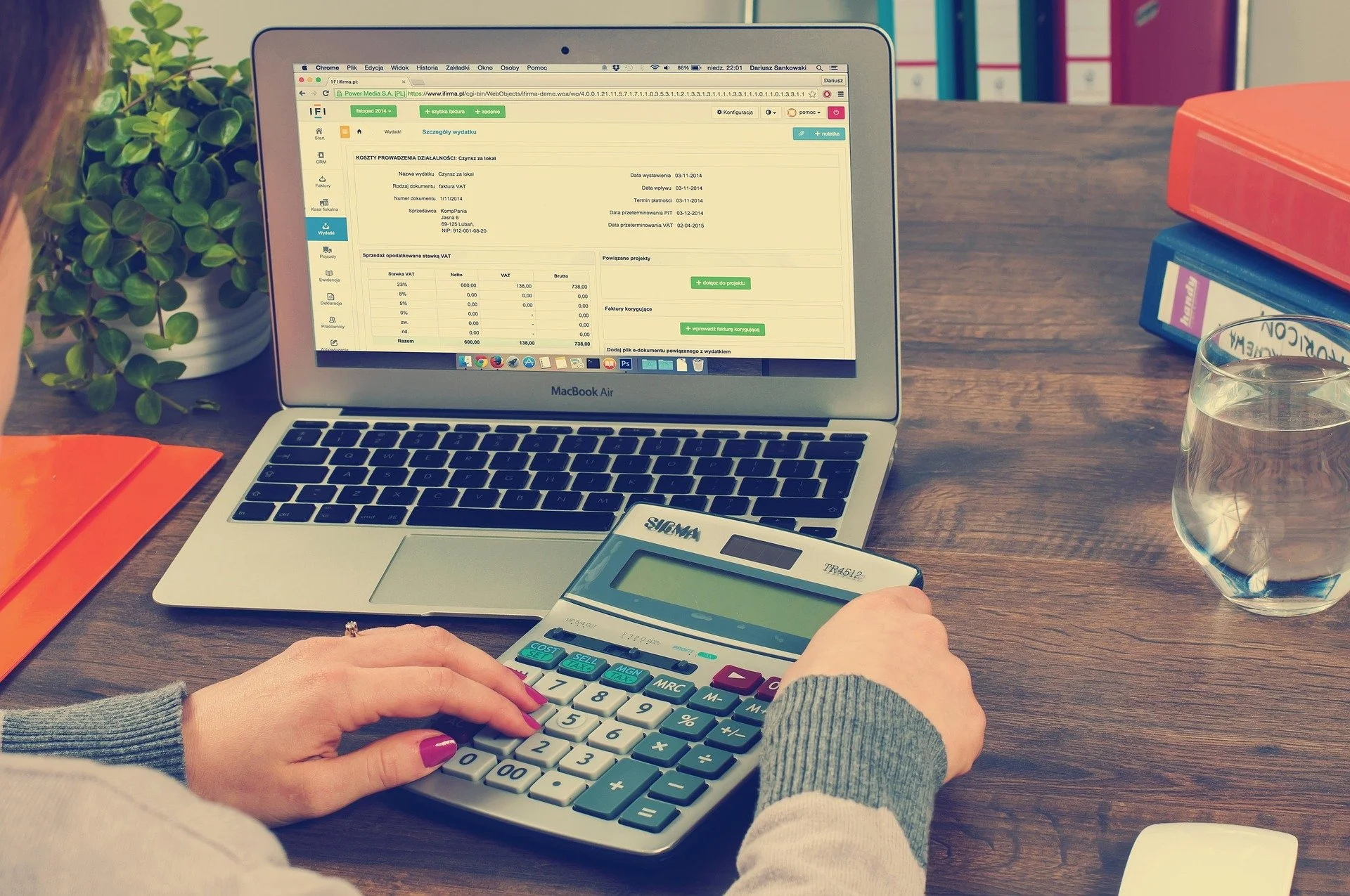Person working at a wooden desk with a laptop, calculator, glass of water, and binders.