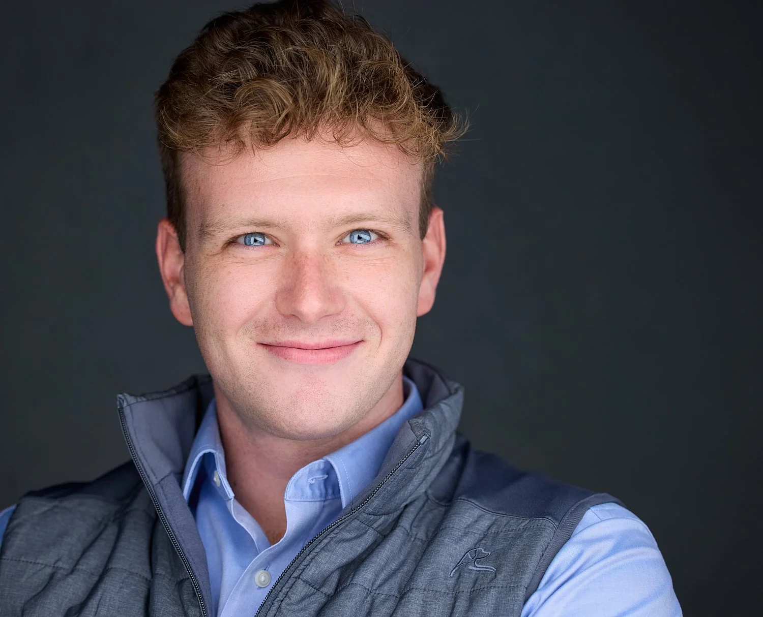 Portrait of a young man with blue eyes, light brown curly hair, wearing a blue collared shirt and a gray vest, smiling against a dark background.