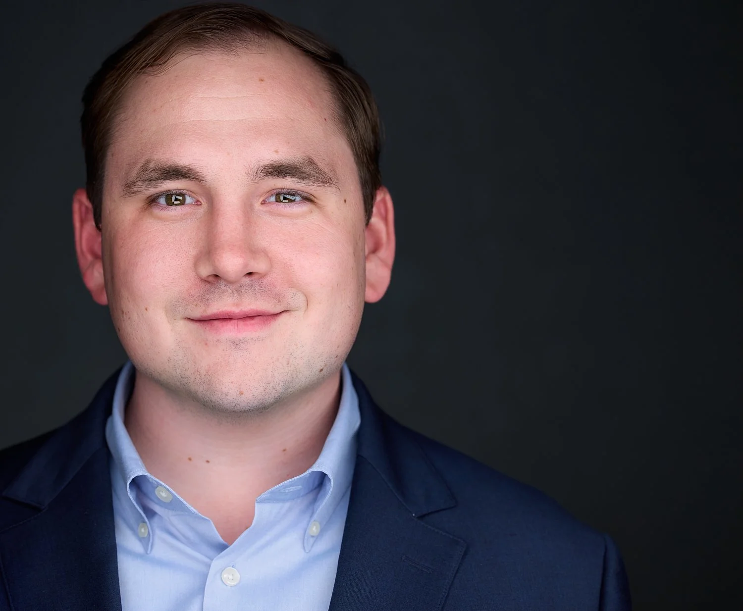 Portrait of a man wearing a dark blazer and a light blue collared shirt, smiling with a dark background.