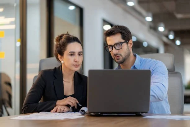 Two professionals, a woman and a man, sitting at a conference table looking at a laptop in a modern office setting.