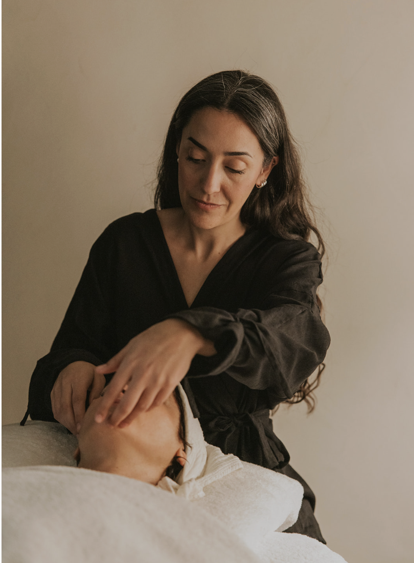 A woman preparing to give a facial treatment to a person lying down with face covered by a towel.