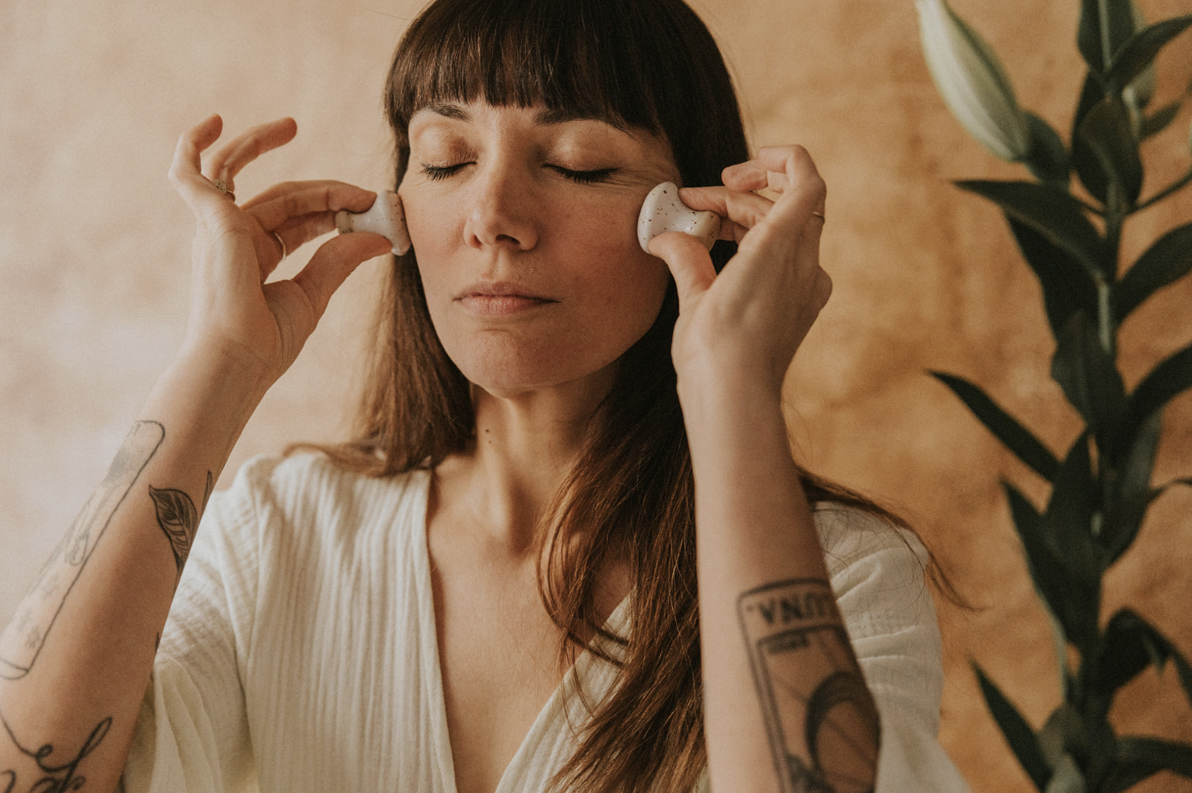 A woman with long brown hair and tattoos on her arms is using facial massage tools on her cheeks with her eyes closed against a warm brown background with green leaves.