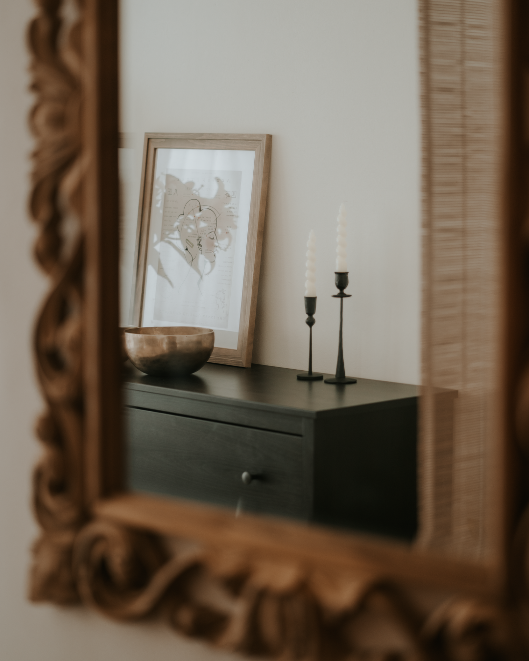 Decorative scene reflected in a mirror with a black dresser, two black candle holders with white candles, a framed outline art print, and a wooden bowl.